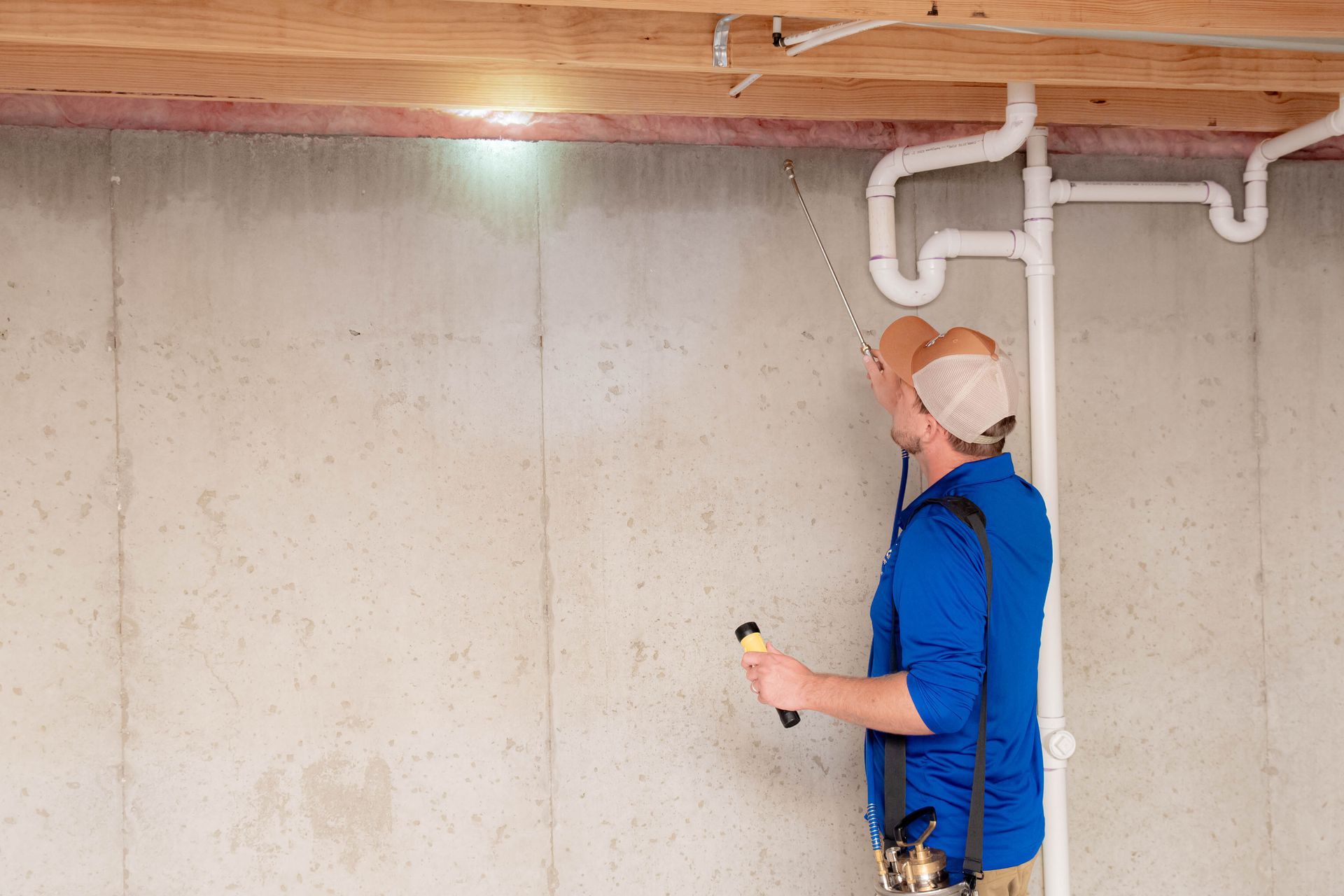 A professional inspecting a basement concrete wall with a flashlight and a telescopic tool near exposed plumbing.