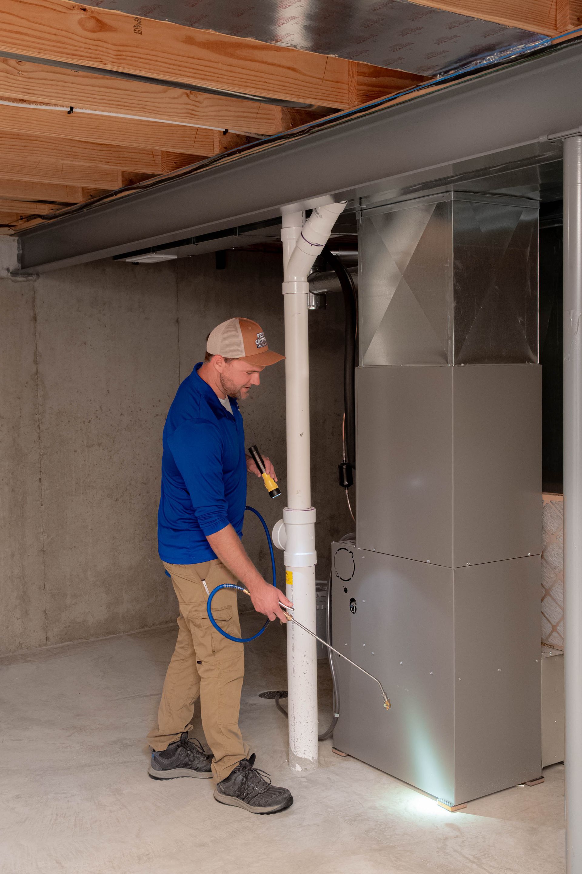 A person in a blue shirt inspects a grey furnace in a basement, pointing a light at the base of the unit.