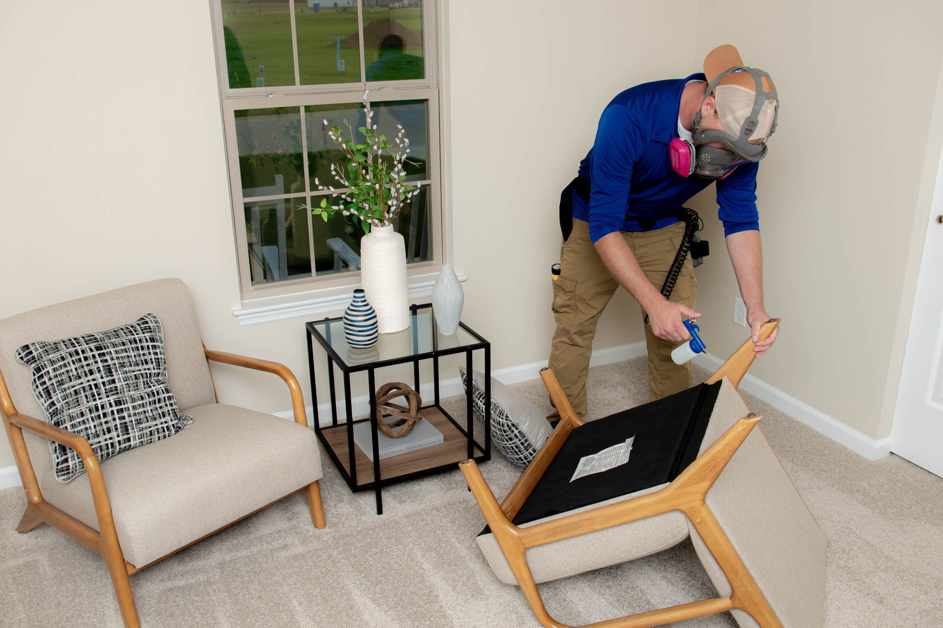 A person wearing a respirator mask sprays furniture in a bright room with a window, side table, and armchair.