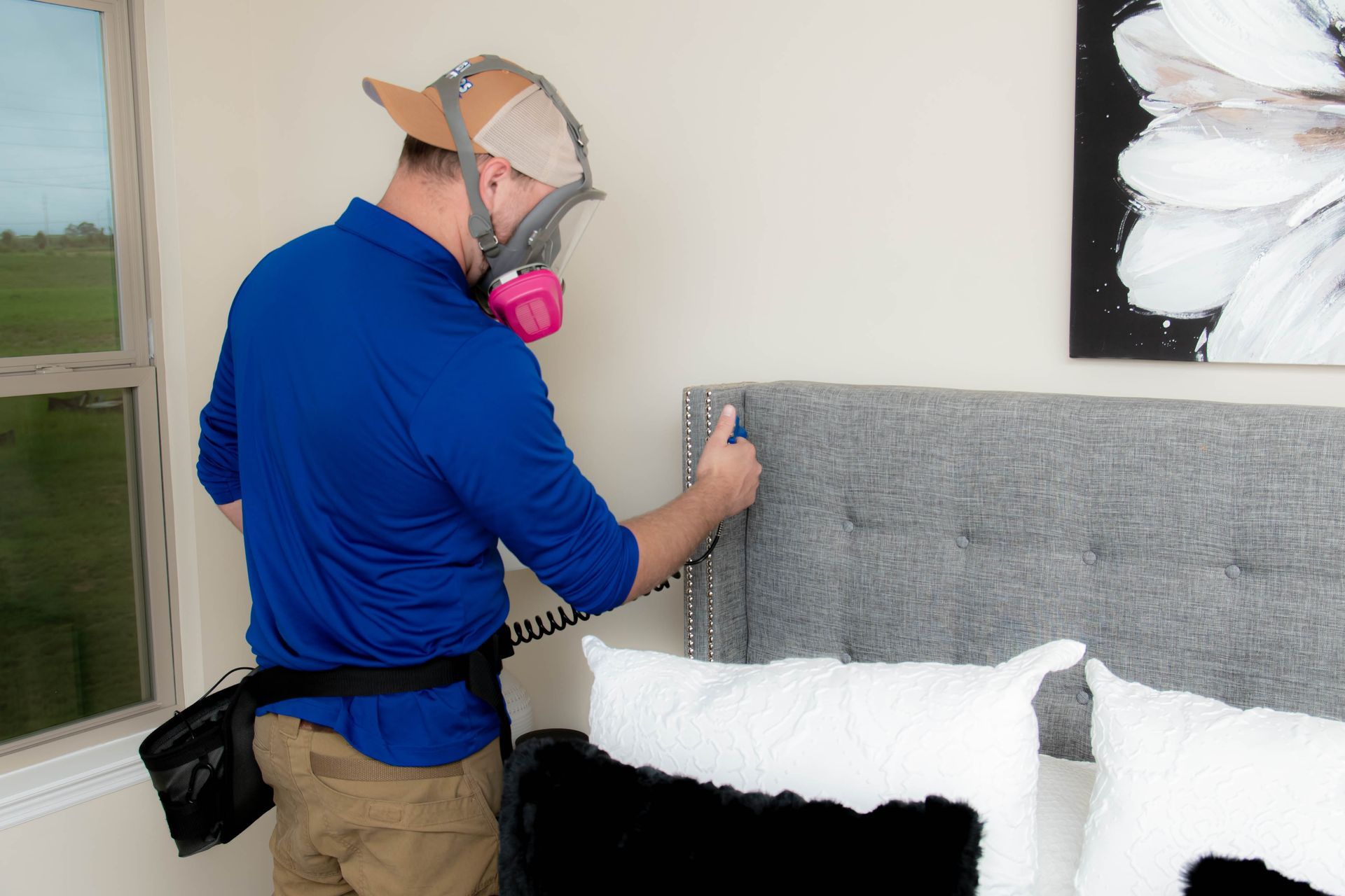 A person wearing a respirator mask and cap cleans a grey tufted headboard in a bedroom.