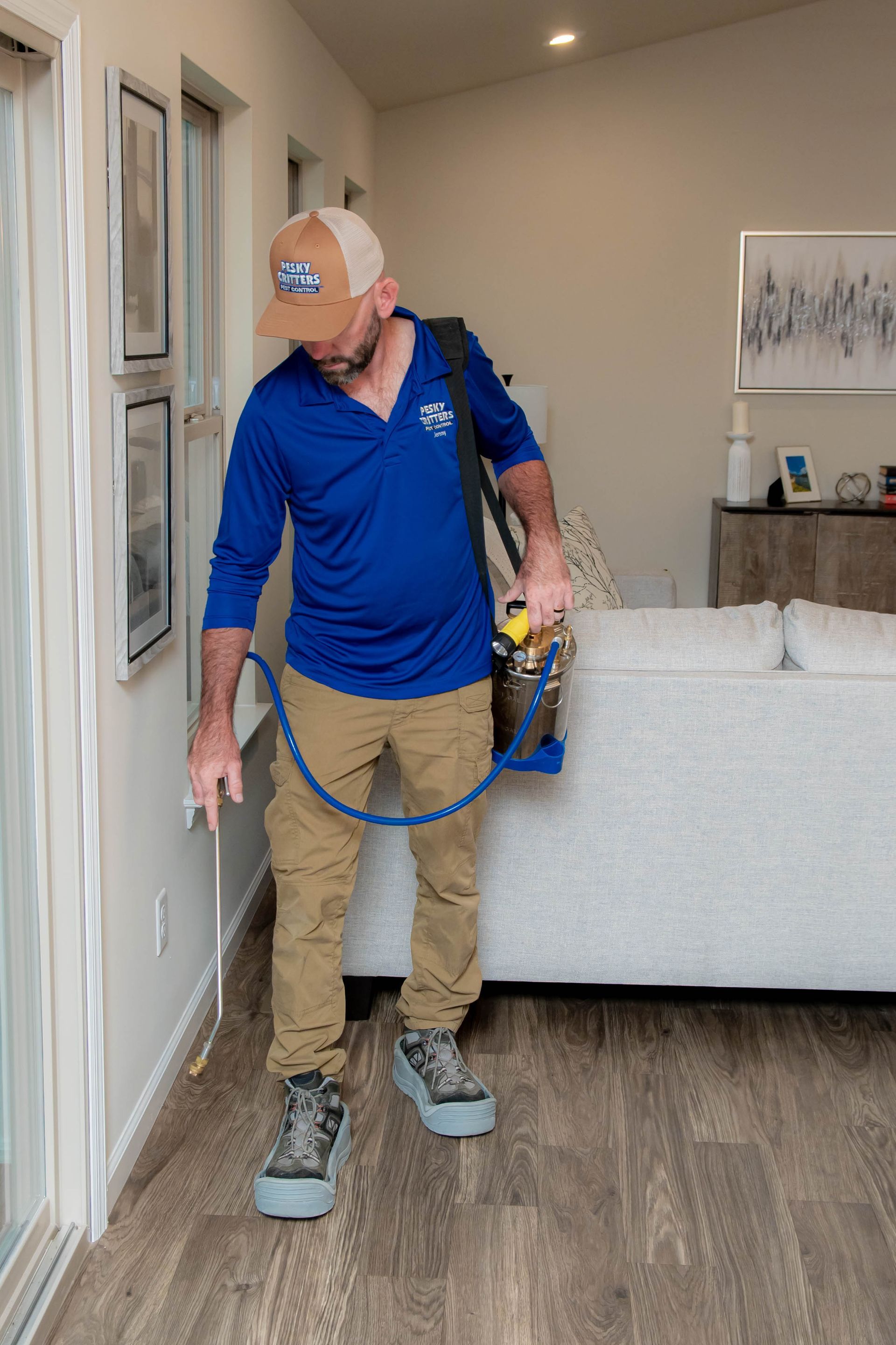 A pest control technician in a blue polo and tan pants sprays a residential interior baseboard with a handheld sprayer.