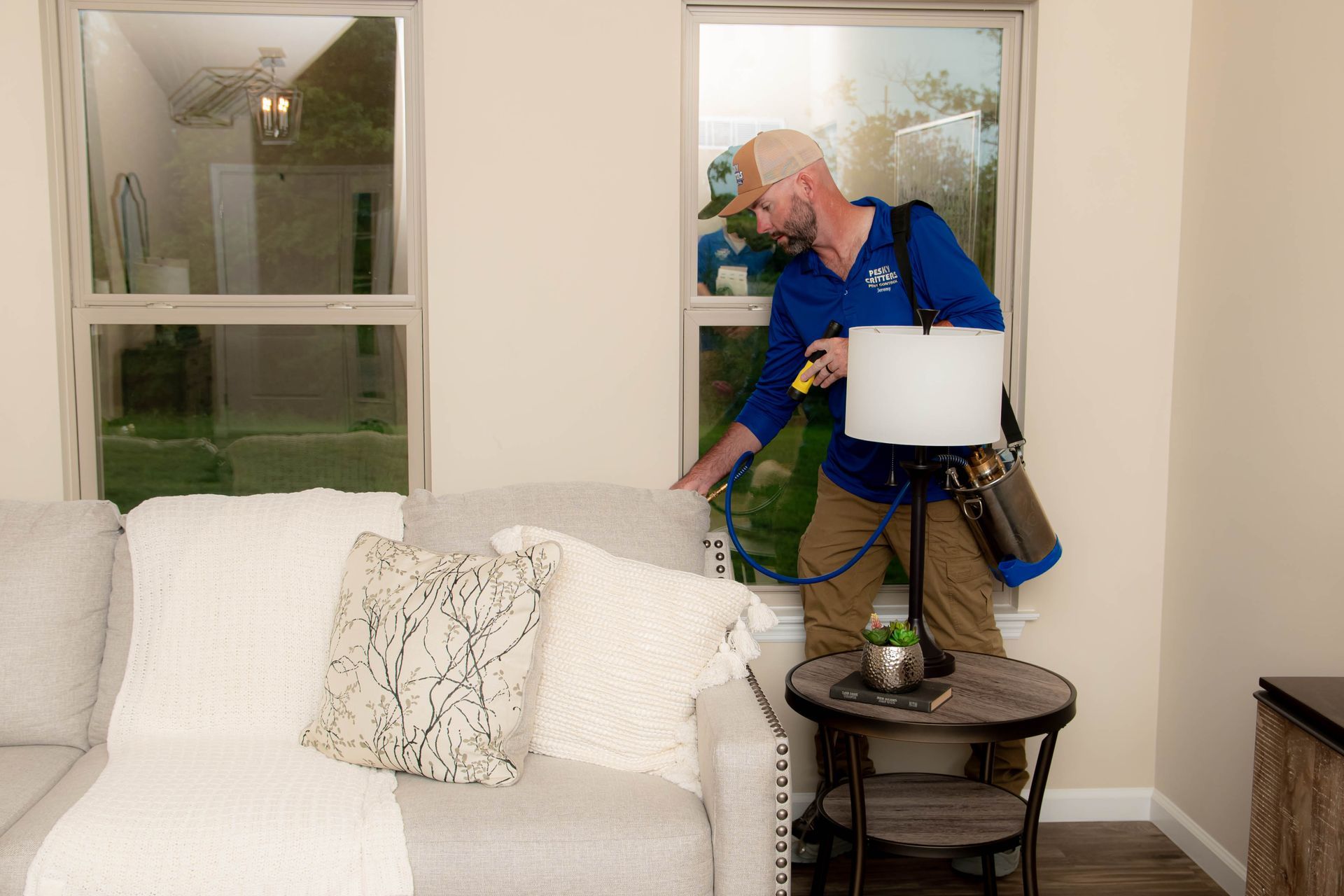A technician in a blue uniform sprays insecticide along the window frame in a living room behind a gray couch.