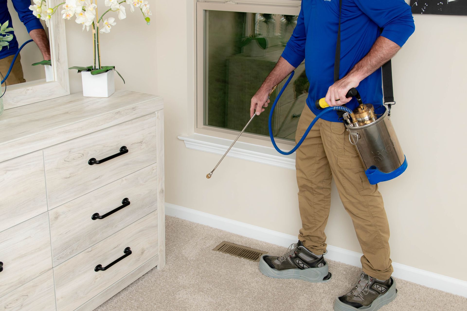 A technician in a blue shirt sprays a perimeter treatment with a metal tank sprayer inside a furnished room.