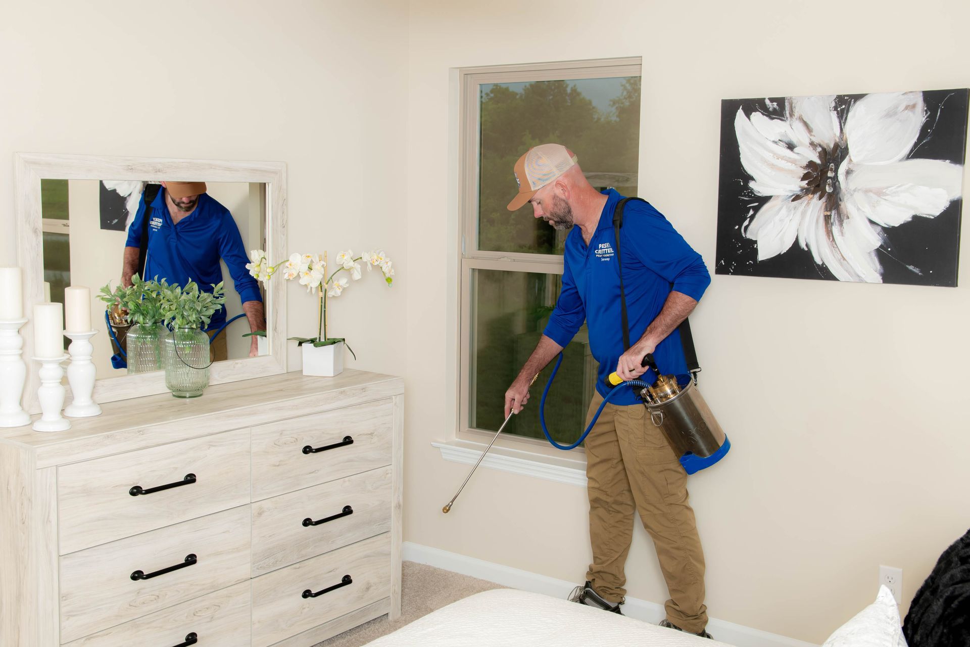 A pest control technician in a blue shirt sprays along a window frame in a bedroom with a dresser and floral art.