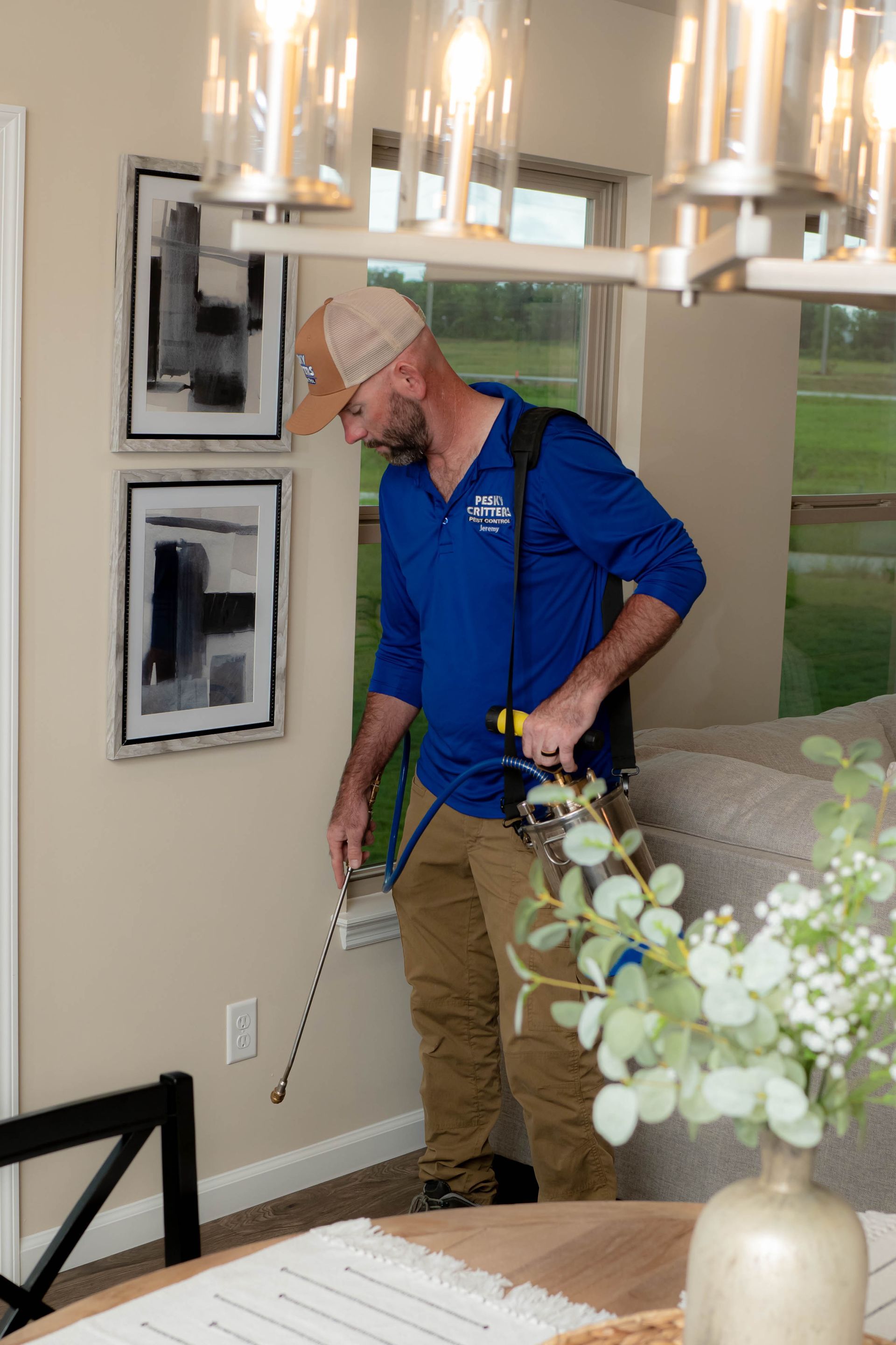 A service technician in a blue shirt sprays insecticide along the baseboards of a residential home.