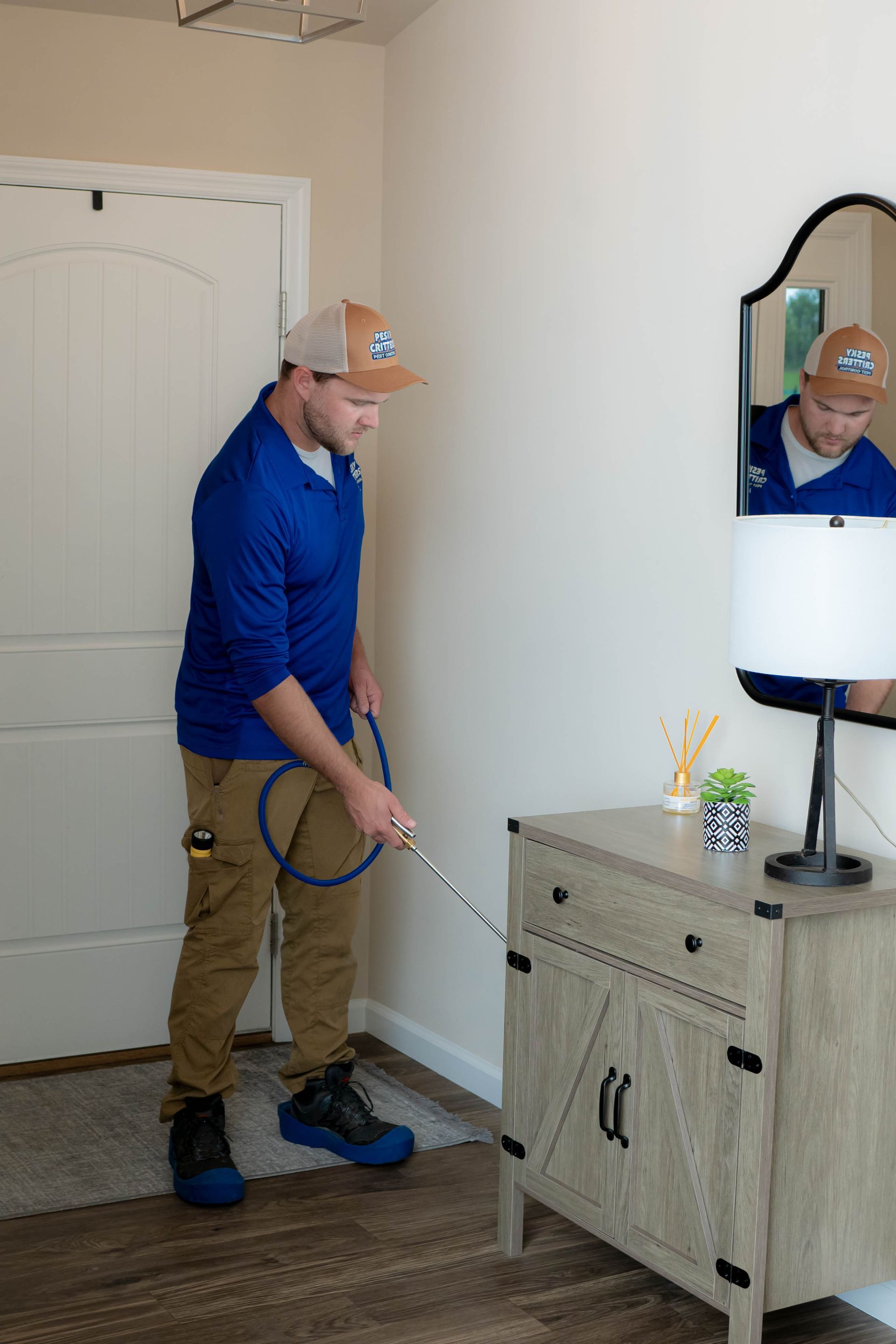 A person in a blue shirt and tan pants sprays a cleaning or pest control solution near a wooden cabinet in a hallway.