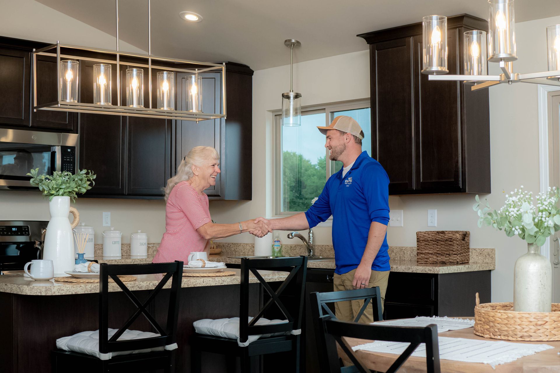 A person in a blue shirt shakes hands with an older person in a kitchen with dark cabinets and a granite countertop.