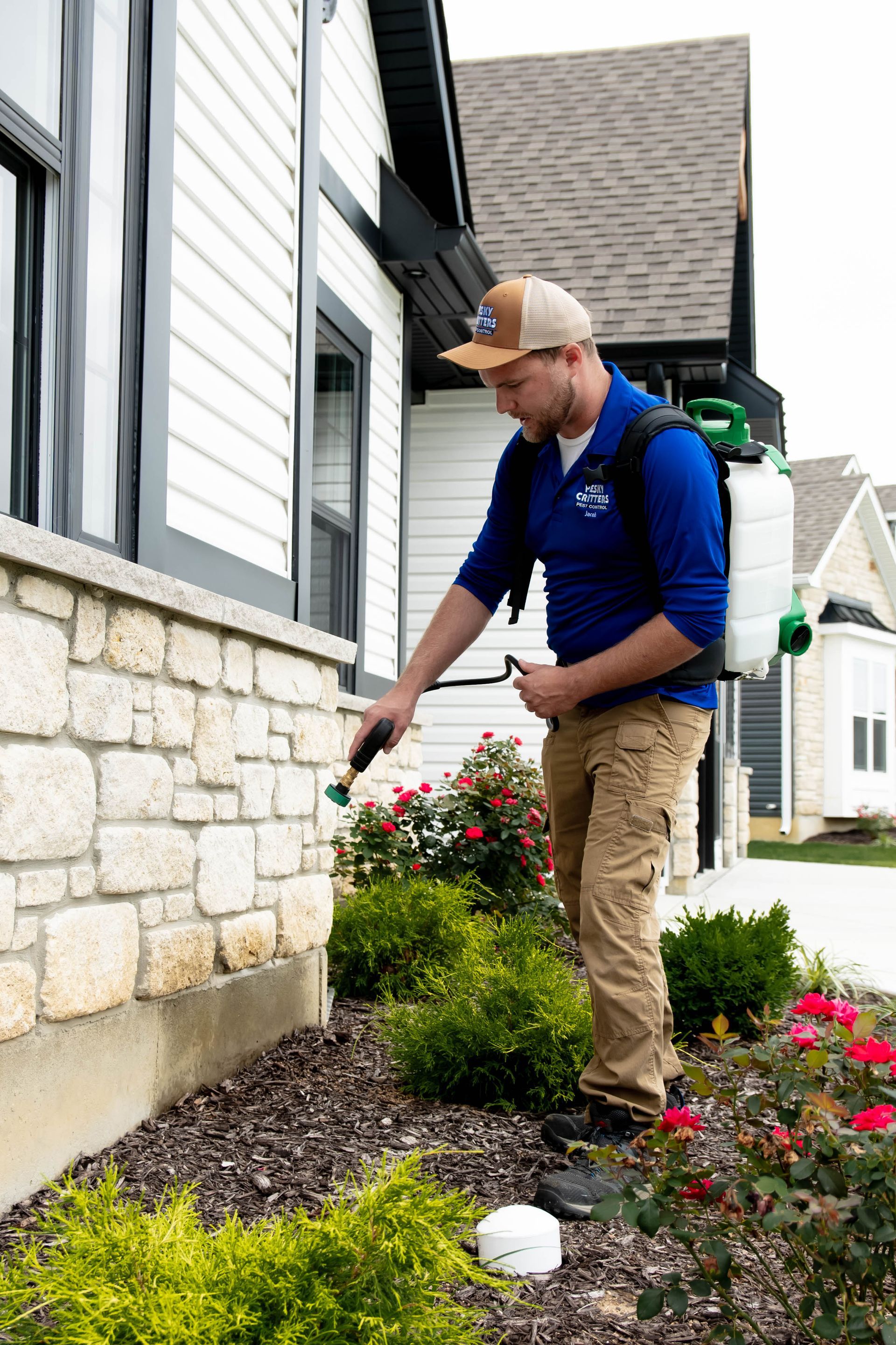 A pest control technician in a blue shirt sprays the foundation of a house with a backpack sprayer near landscaping.
