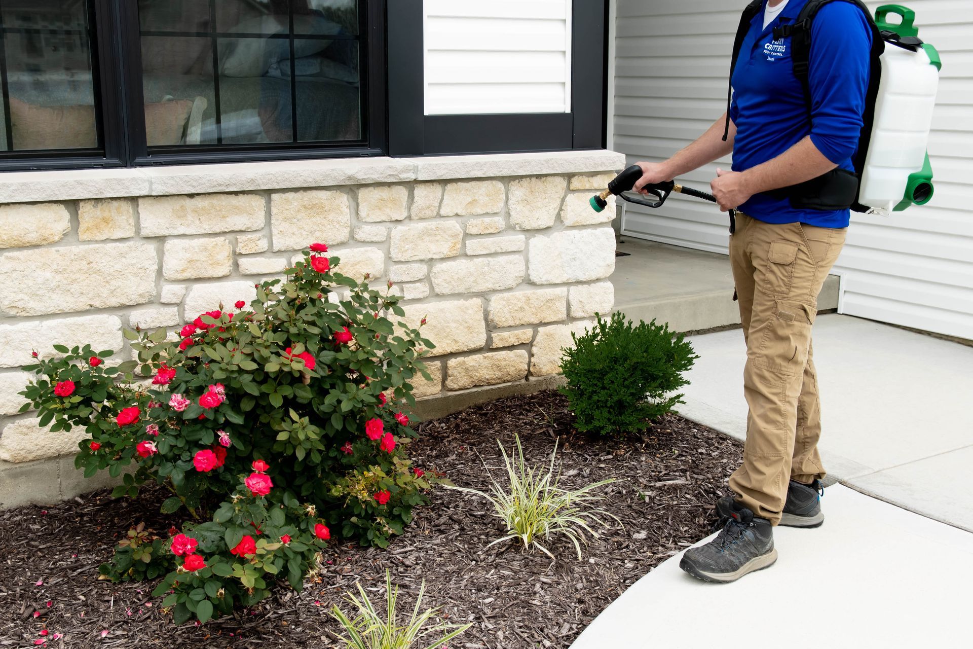A service technician in a blue uniform applies pest control spray to the stone foundation of a house.