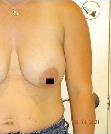 Torso with breasts; one nipple obscured. Wears jeans, jewelry. Tan skin, indoor setting, close-up shot.