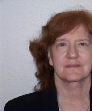 Woman with reddish hair, wearing a black blazer, looking forward. White wall background.