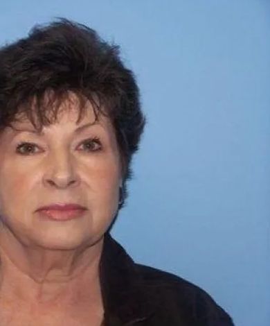 Woman with short dark hair, wearing a black top, against a blue background, looking at the camera.