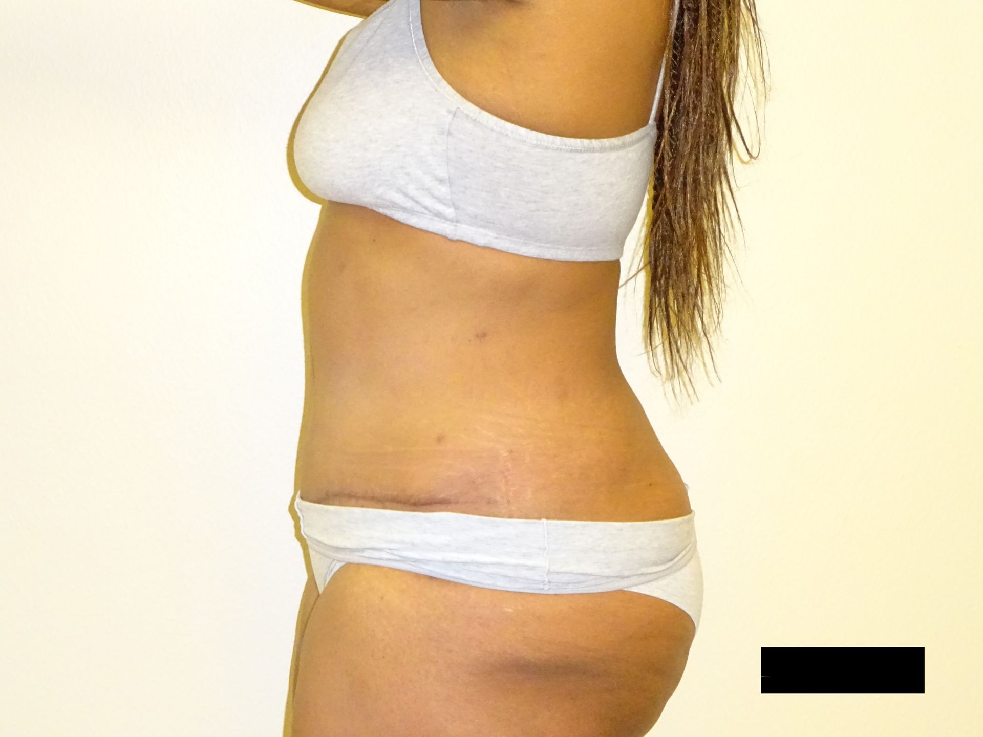 Woman in white underwear and bra, standing sideways, in front of a white wall.