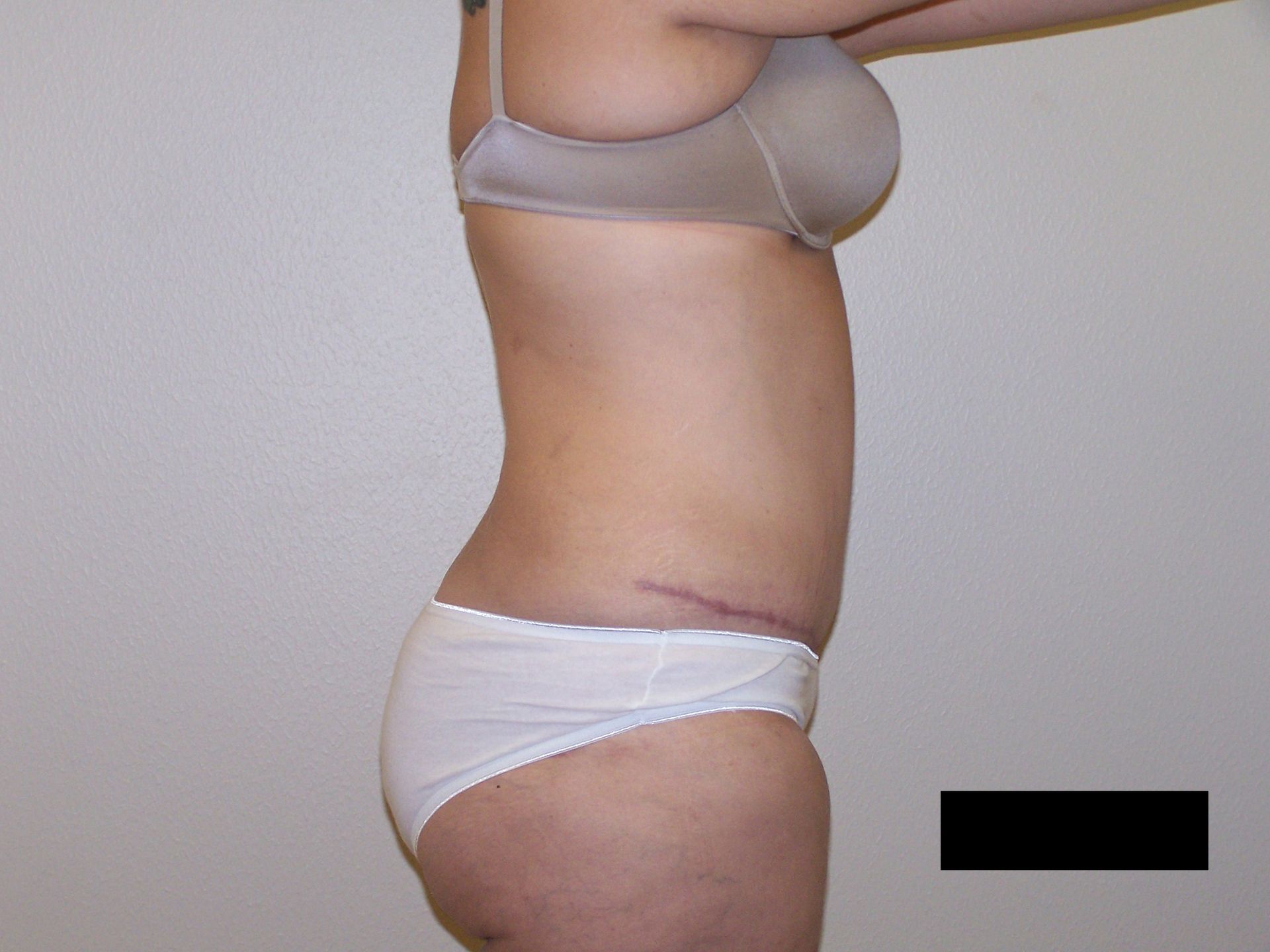 Woman in white underwear and bra, standing sideways, in front of a white wall.