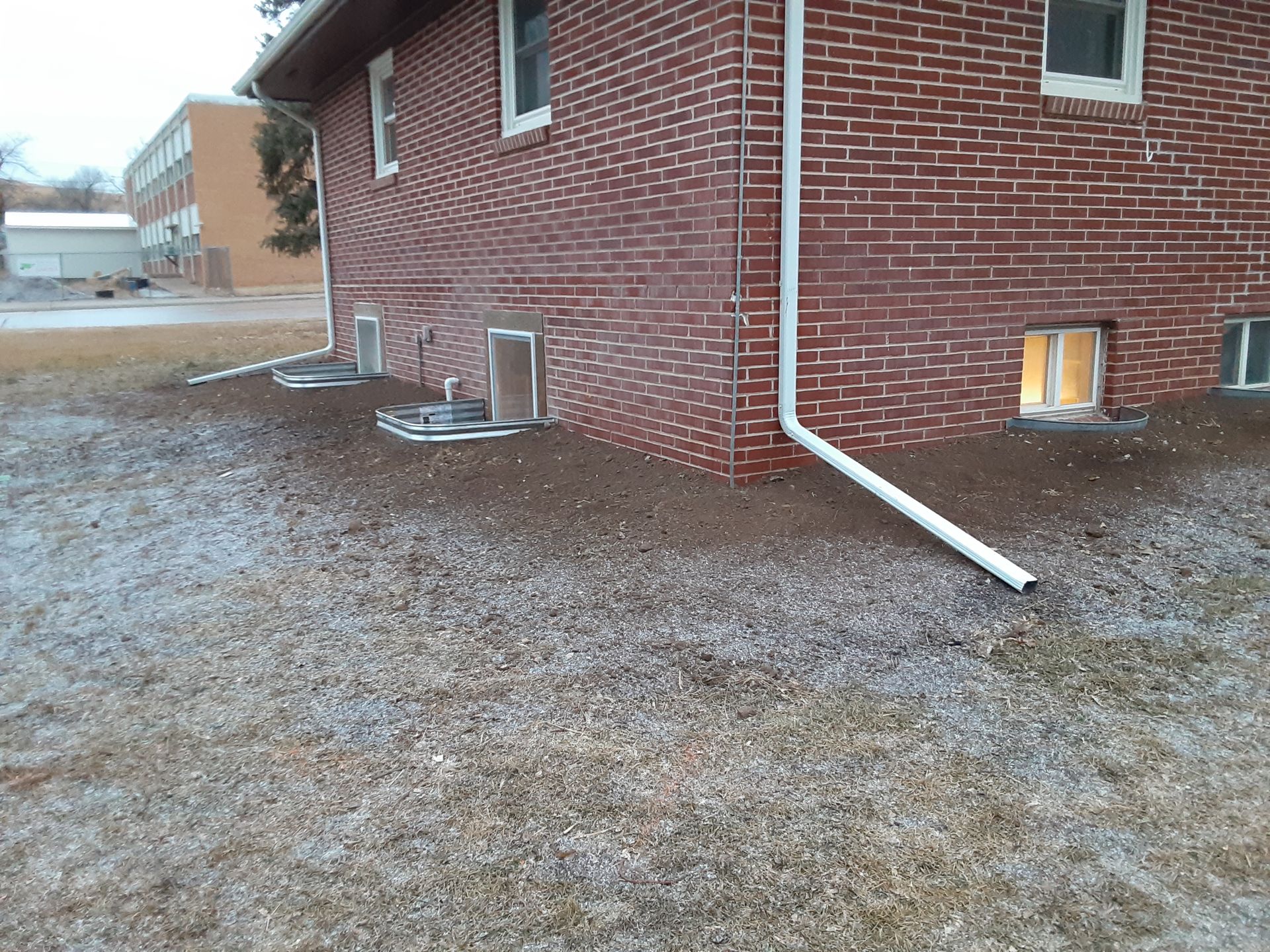 Red brick building corner with small basement windows, gravel and dead grass yard.