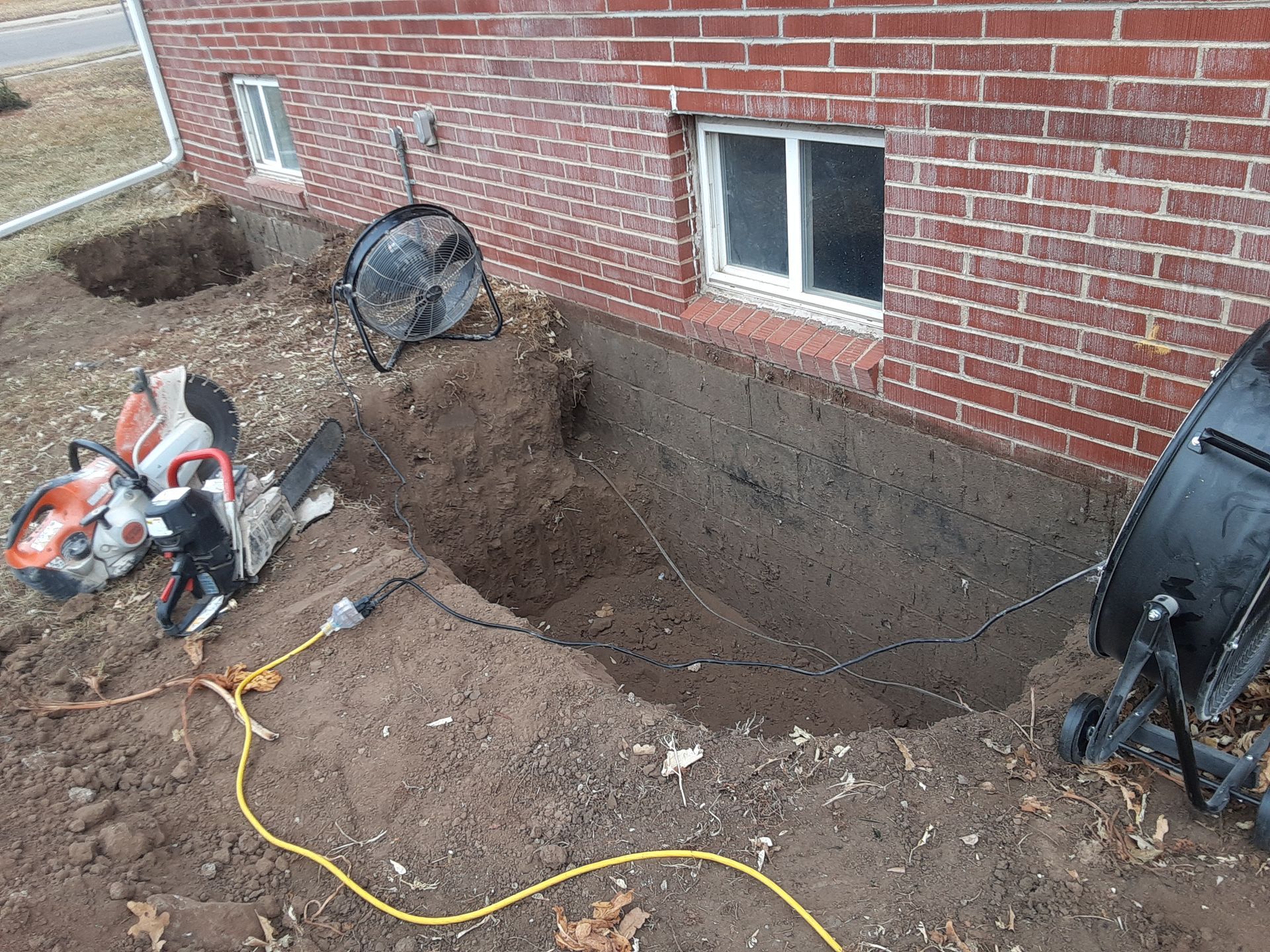 Excavation near a brick building's foundation. A saw, fan, and dirt are visible. A basement window is also present.