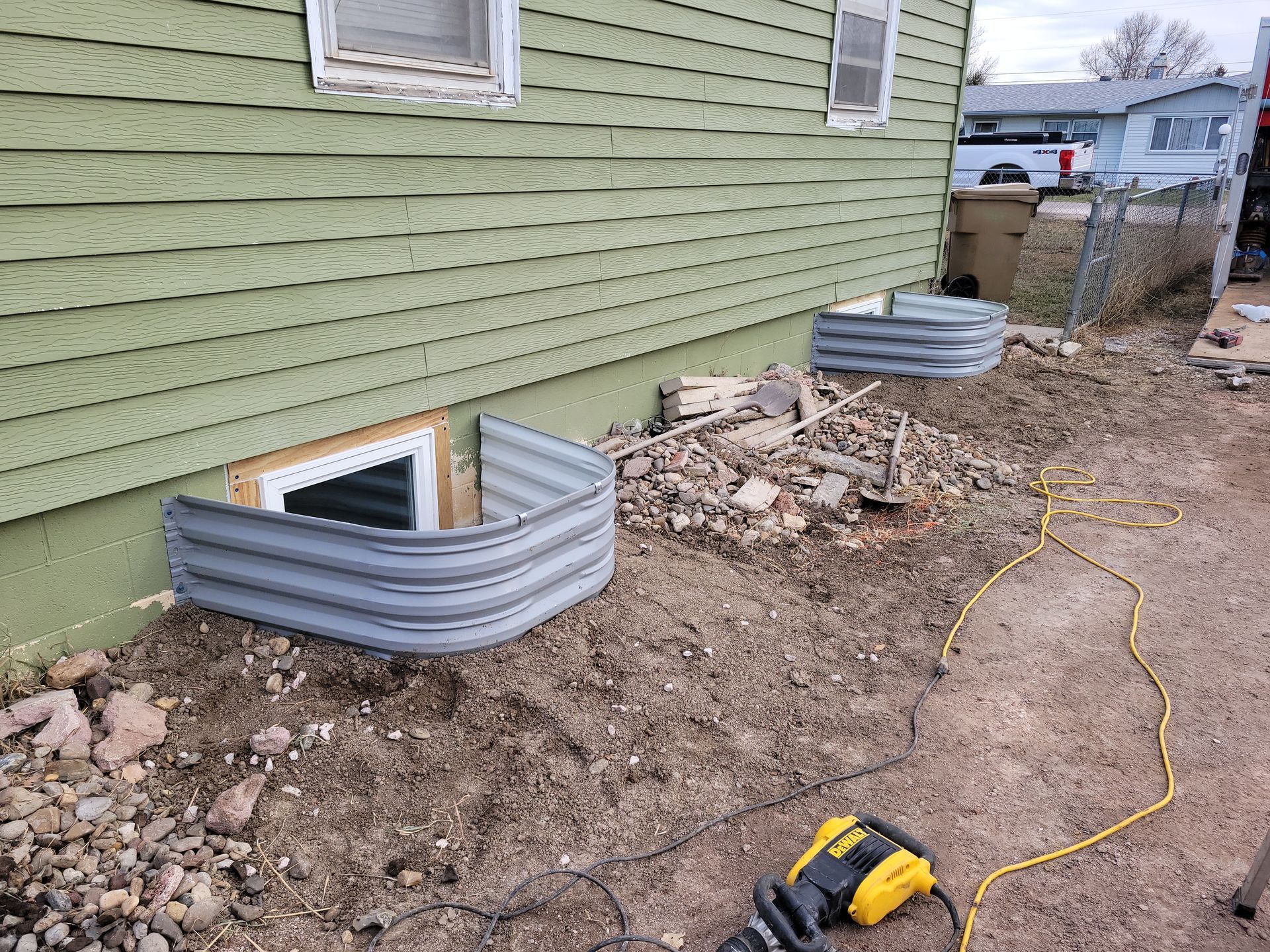 Exterior view of a house with two metal window wells and a dirt yard. A power tool and debris are visible.