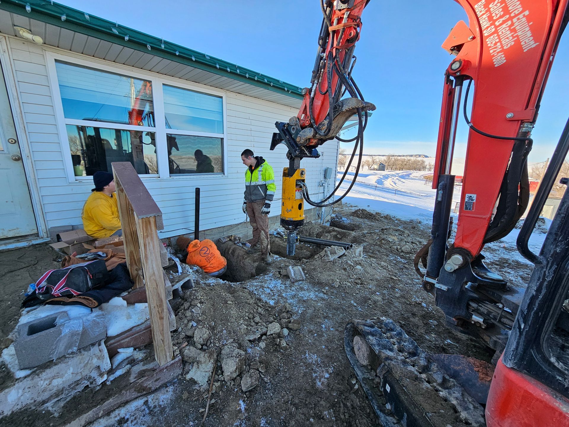 Construction site: excavator drilling near a building; two workers nearby, snowy ground.