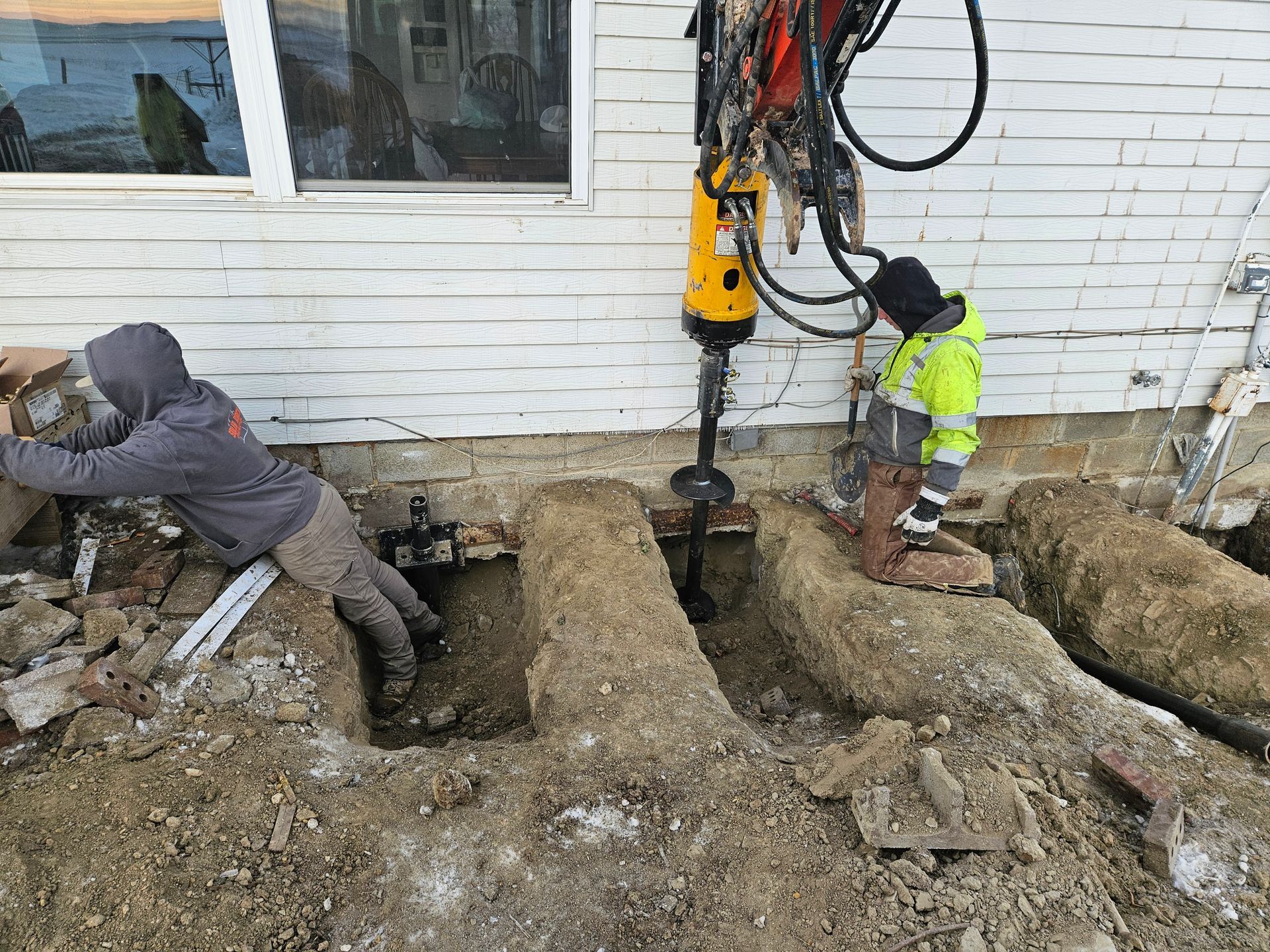 Two workers using equipment to dig near the foundation of a white building.
