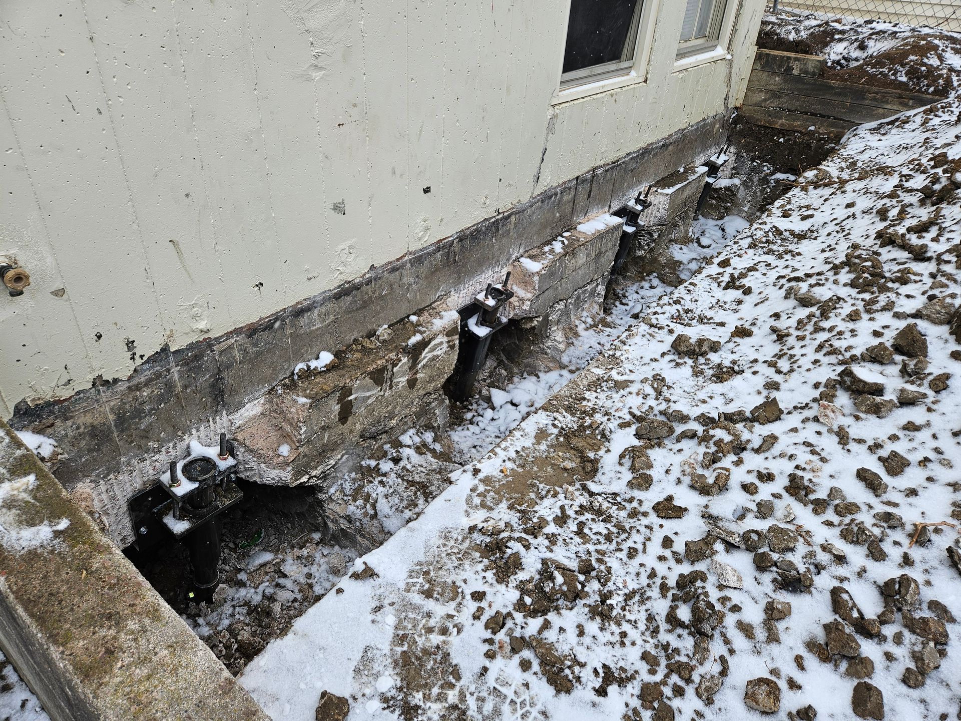 Base of a building with exposed foundation in a trench filled with dirt and snow.