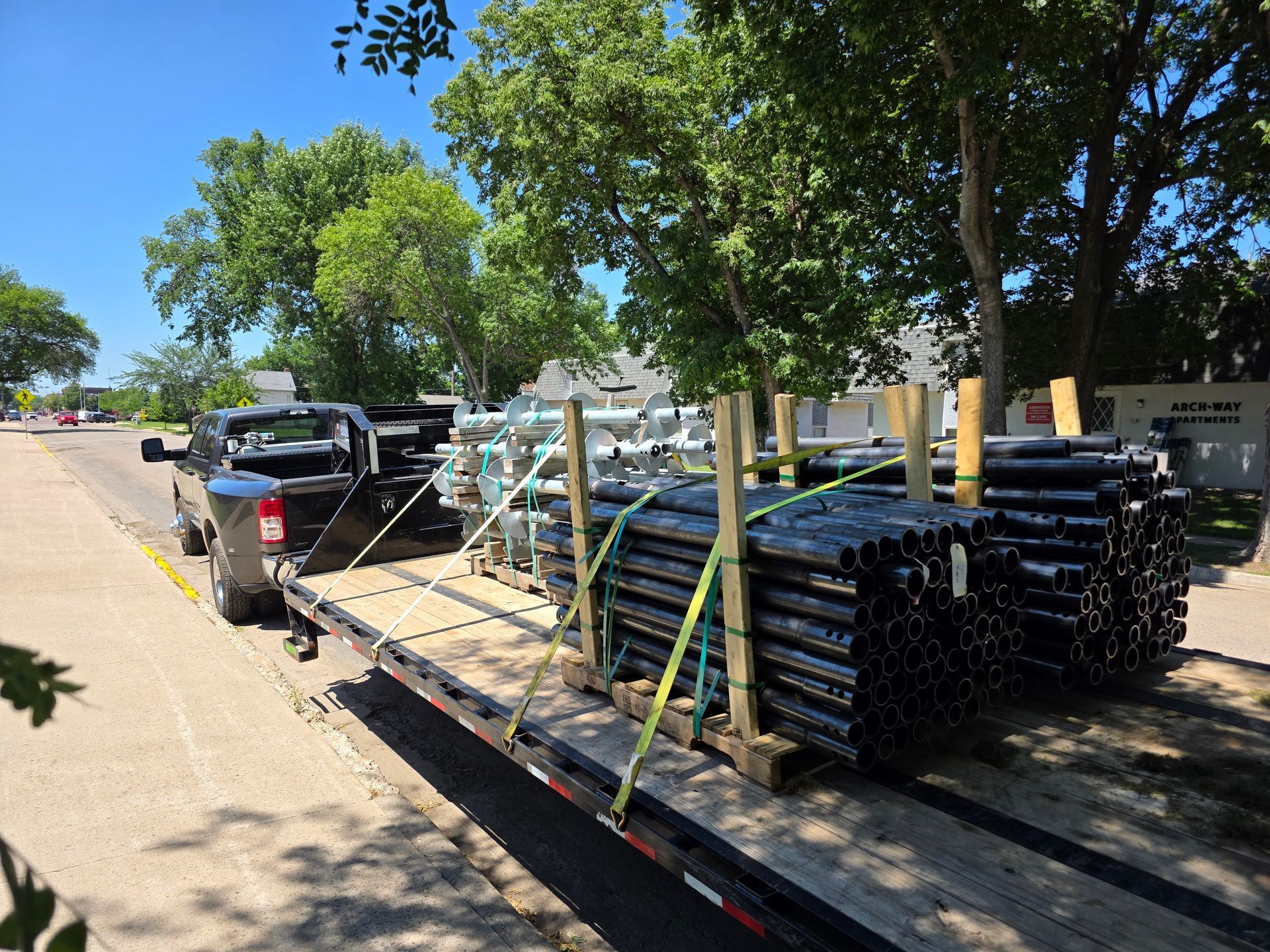 Truck pulling a flatbed trailer loaded with stacks of black pipes, parked on a street near trees.
