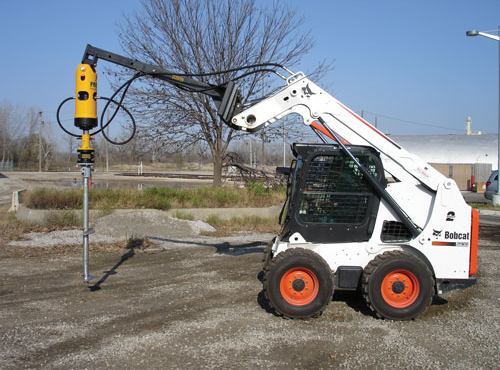 White Bobcat skid-steer with a yellow post driver installing a metal post on gravel.