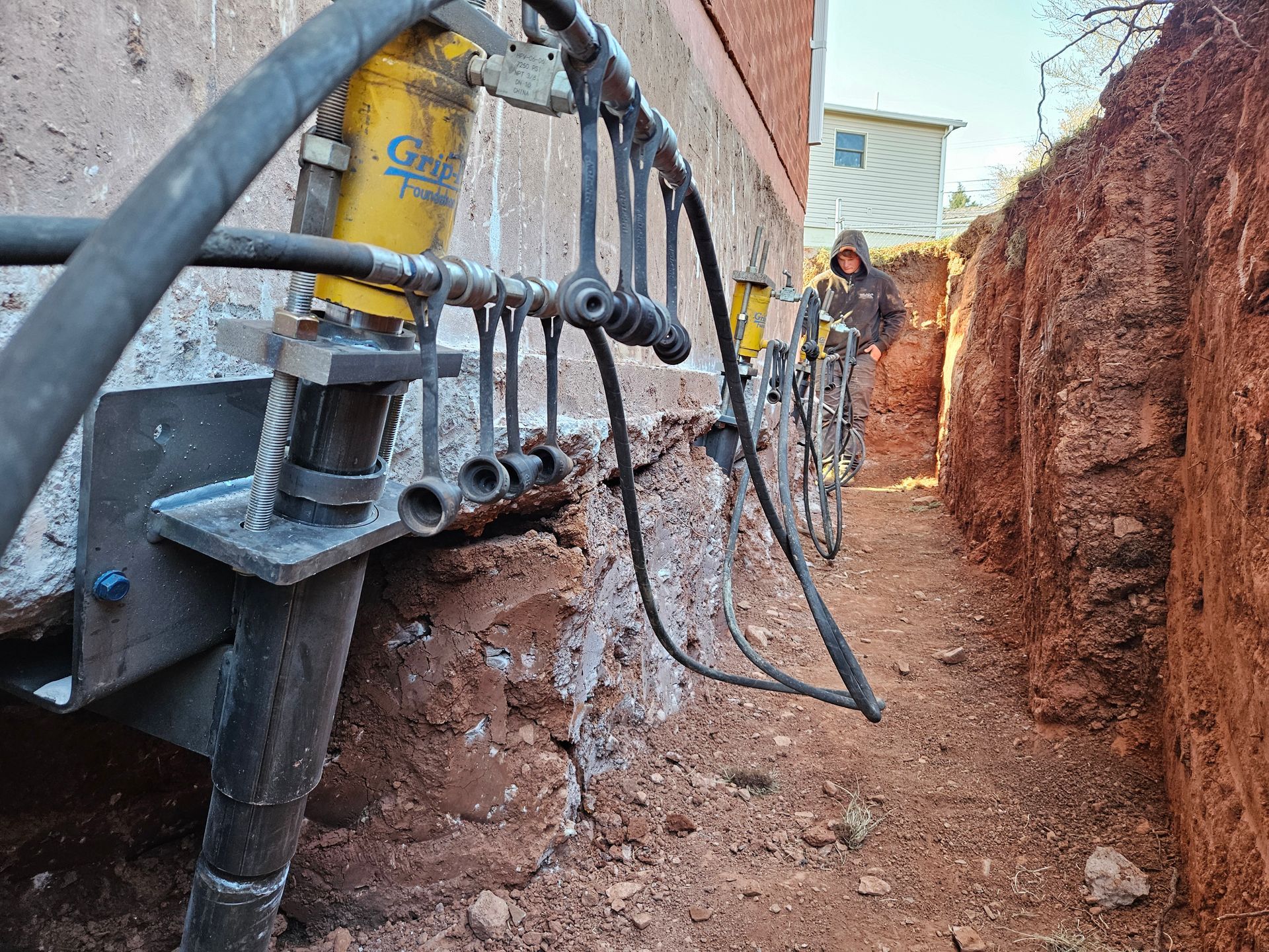 Foundation repair with hydraulic jacks, trench alongside building. Person in background.
