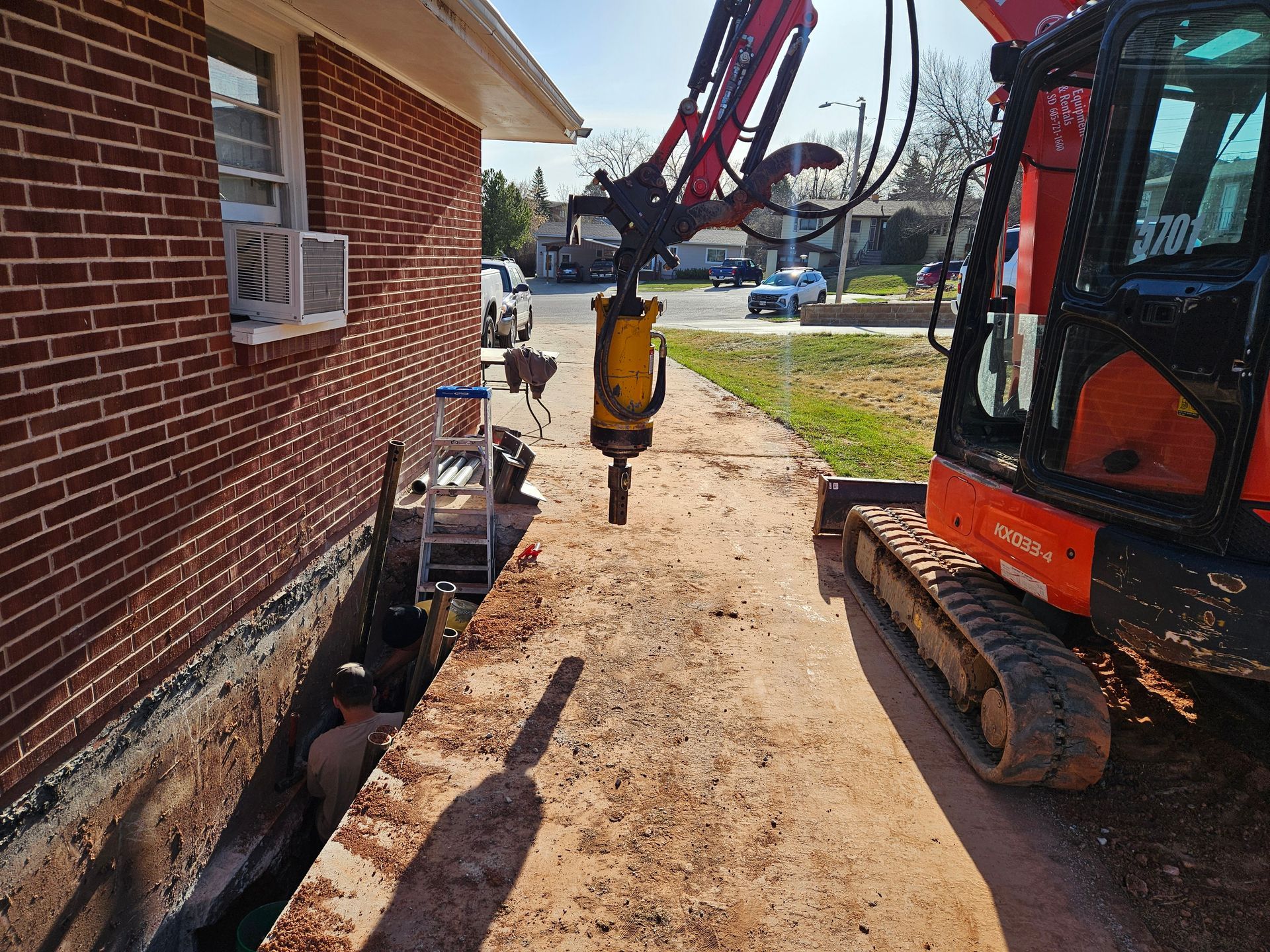 An excavator drilling near a brick building. Trench alongside the building. Sunny day.