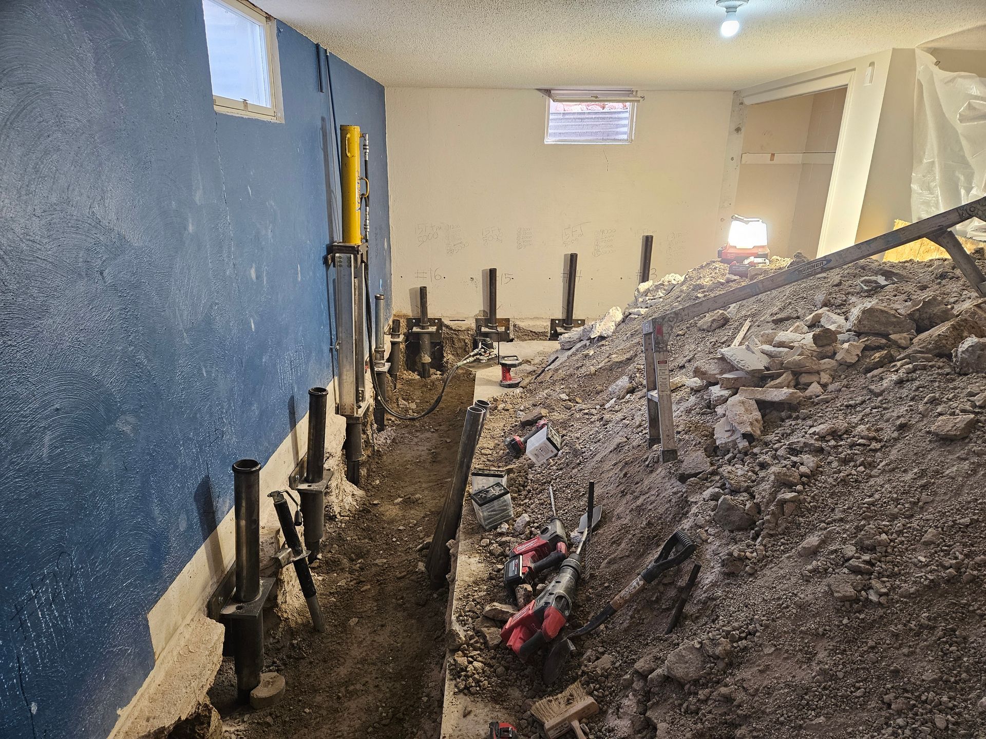 Basement interior under construction, soil trench with support jacks, debris pile, and blue and white walls.
