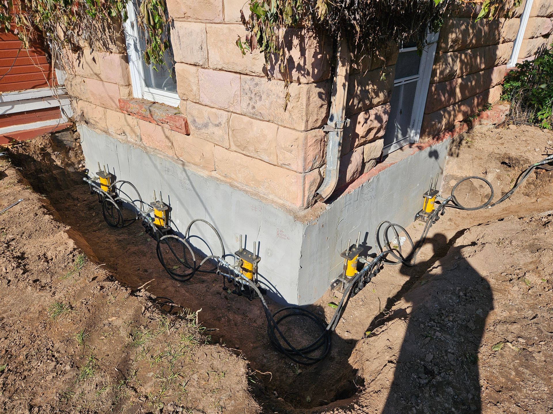 Excavation around a building's foundation. Several yellow jacks are connected with black wiring.