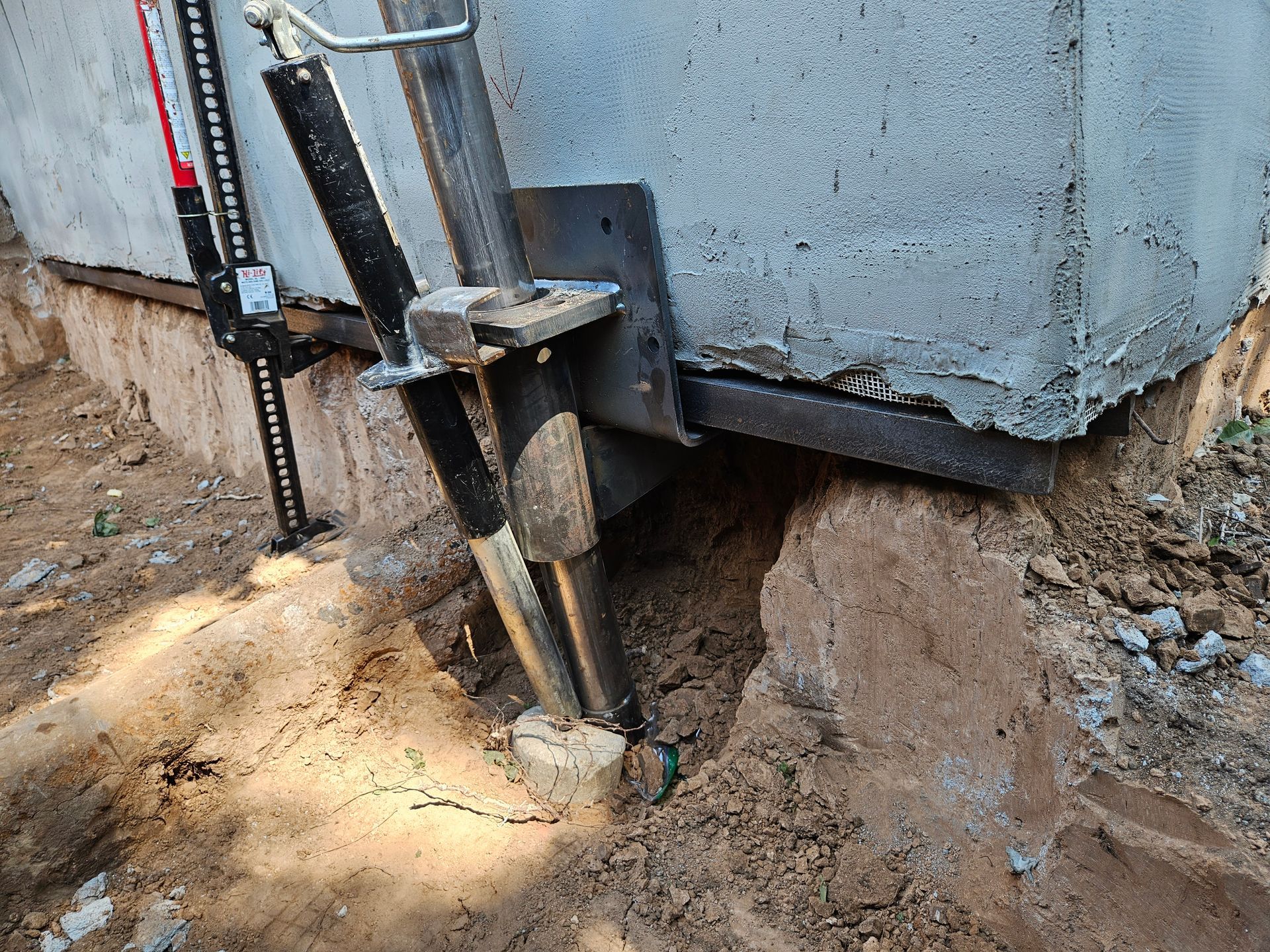 Jack and metal bracket supporting a concrete foundation corner. Soil and dirt surrounding.