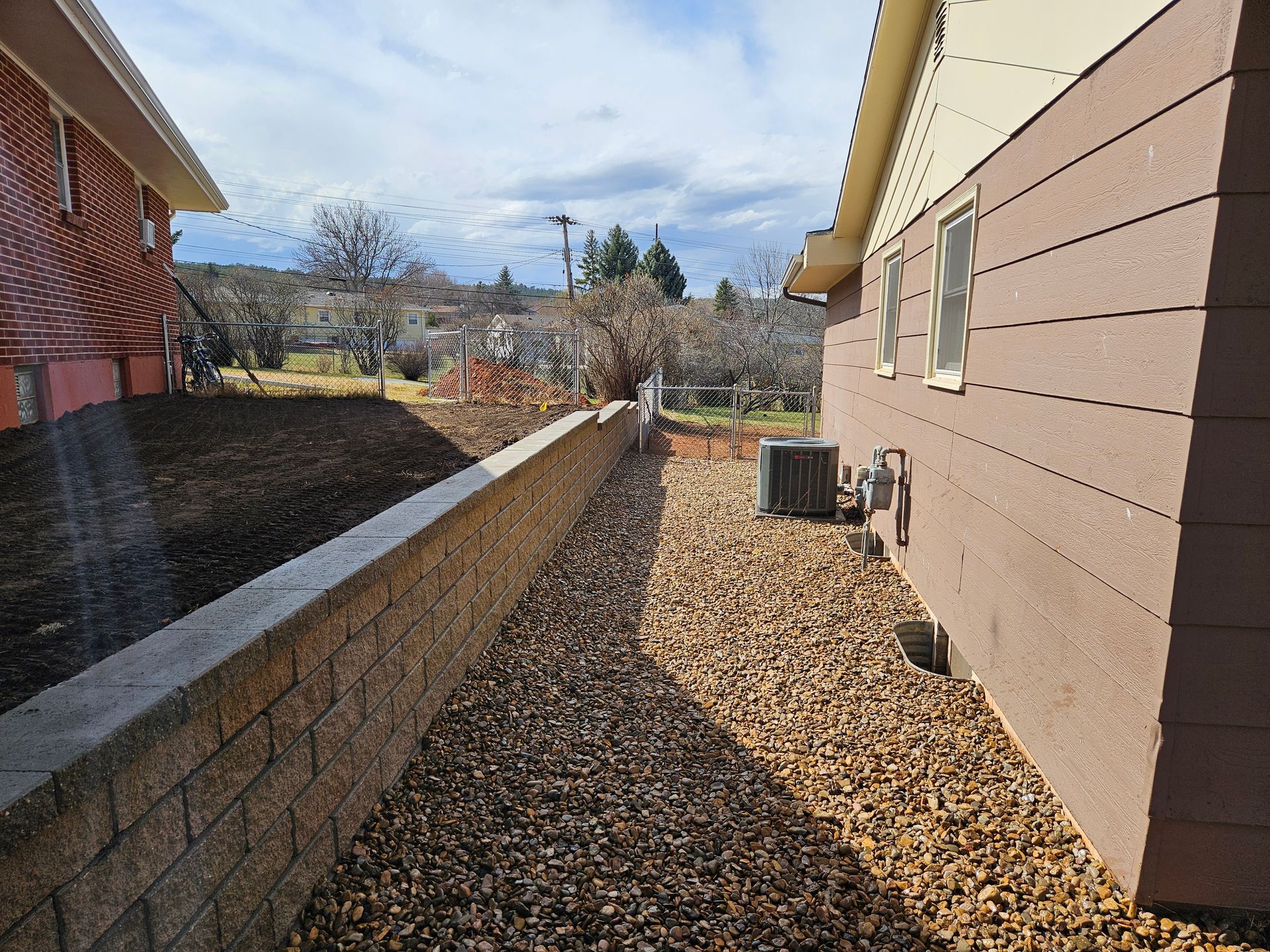 Stone retaining wall along a gravel path next to a brown house with air conditioning unit.