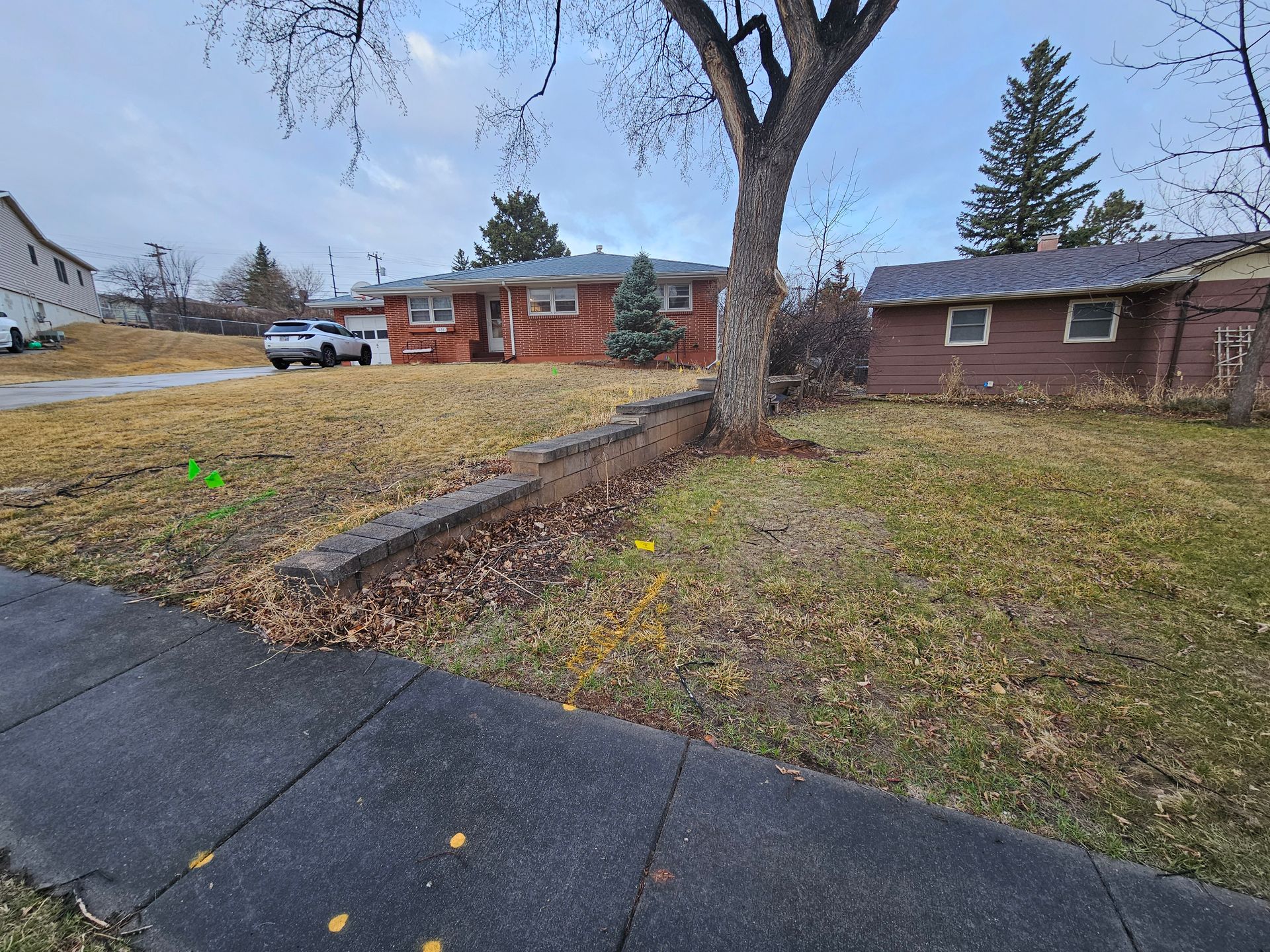 Residential yard with low brick retaining wall, tree, house, sidewalk, and parked car on a cloudy day.