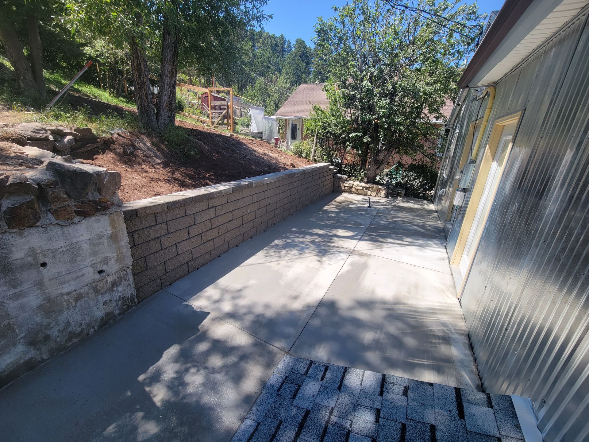 Concrete patio with retaining wall, trees, and corrugated metal building. Sunny day.