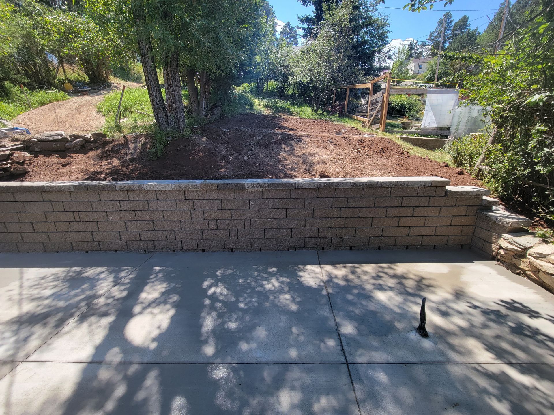 Retaining wall of textured blocks backs a concrete patio, with a dirt hill, trees, and greenery beyond.