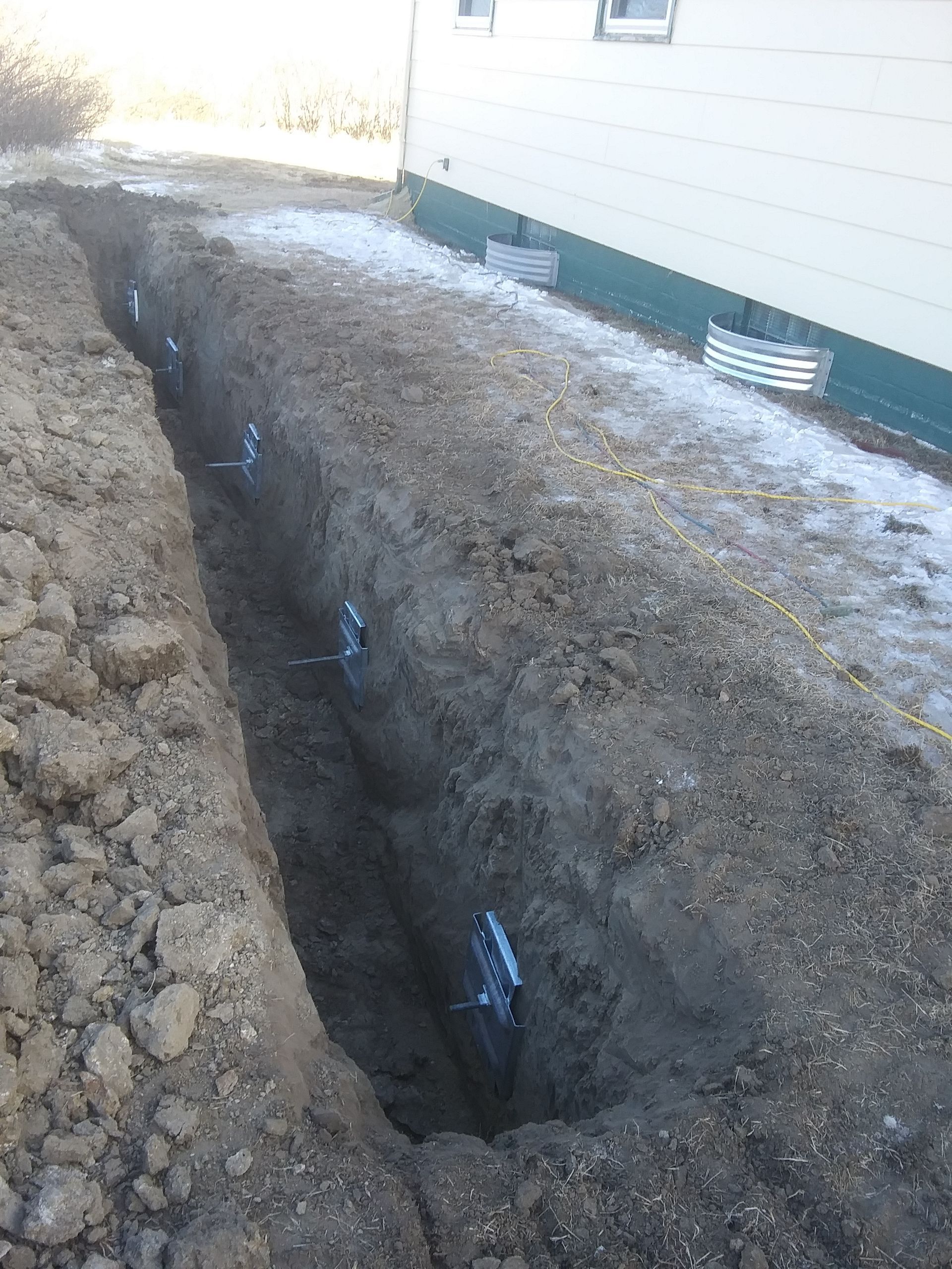 Long trench dug along a building's foundation, containing pipes secured with clips.