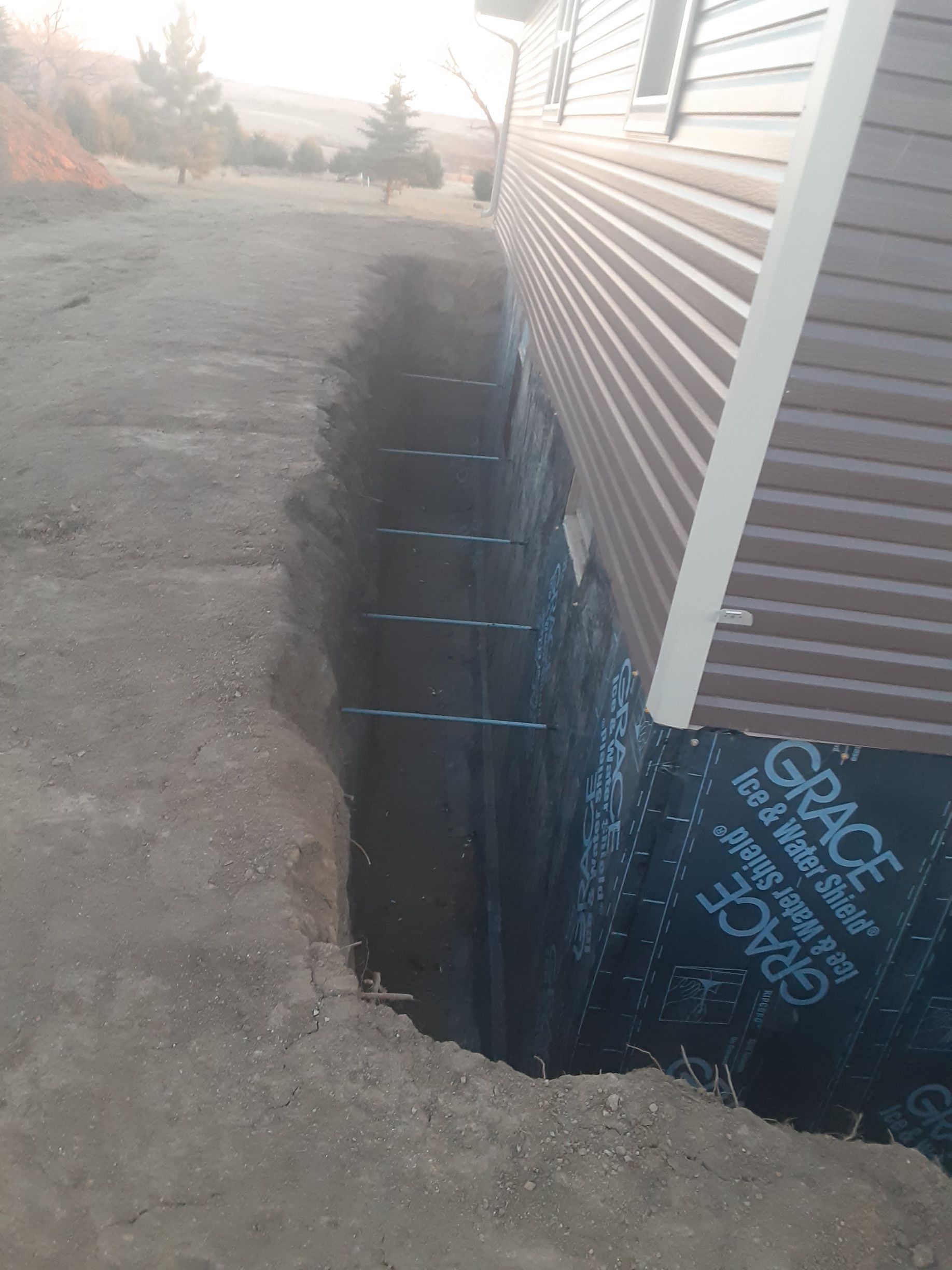 Trench dug next to a building's brown siding.  Black waterproof membrane covers the foundation wall.