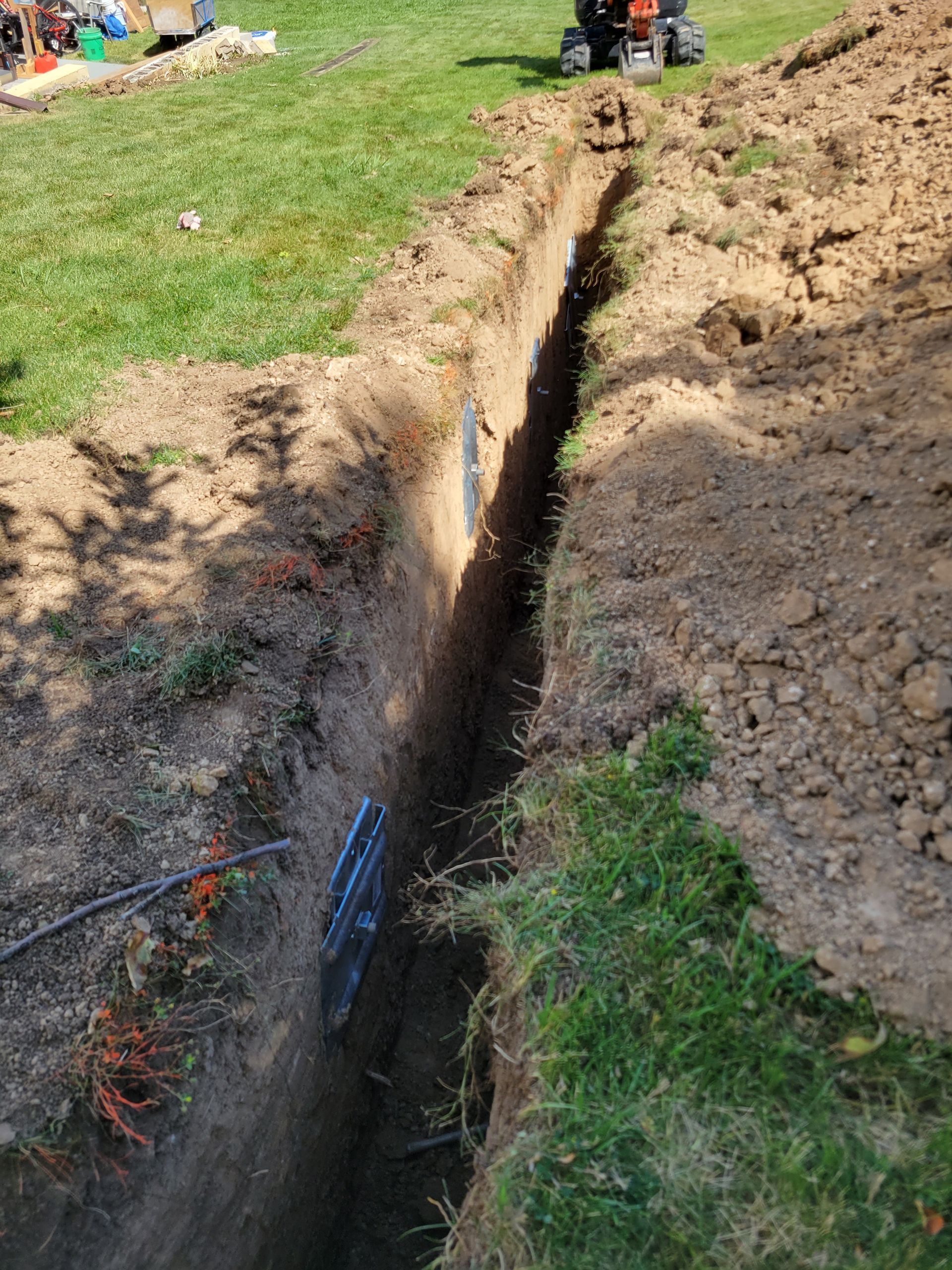A narrow trench dug through a grassy yard, with visible black piping. A small excavator is in the background.