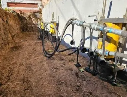 Excavated trench with yellow hydraulic jacks attached to a concrete wall. Black hoses and metal fittings visible.