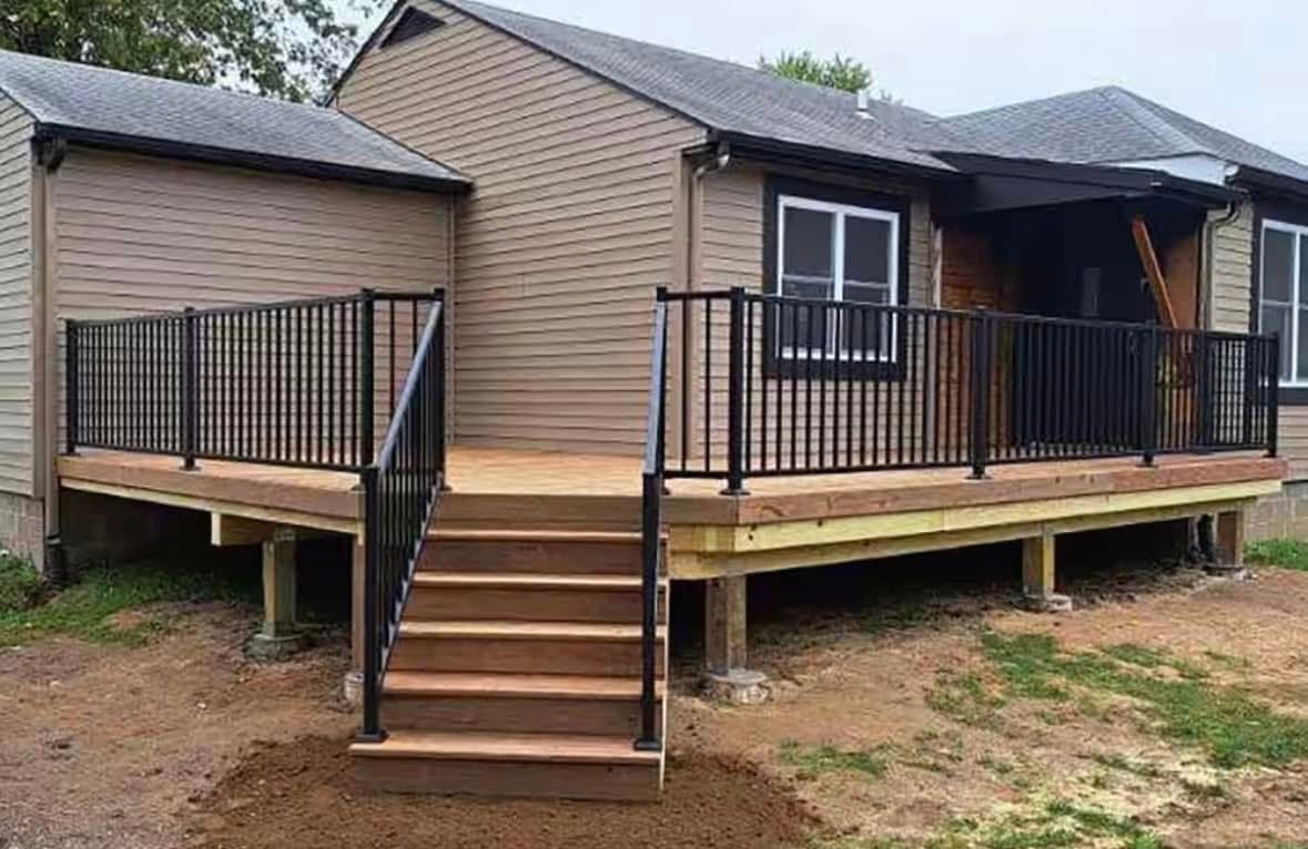 Small wooden house with front porch, stairs, and black railing on bare ground