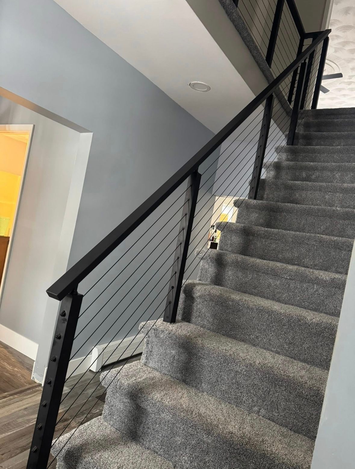 Carpeted indoor staircase with black railing beside a gray wall.