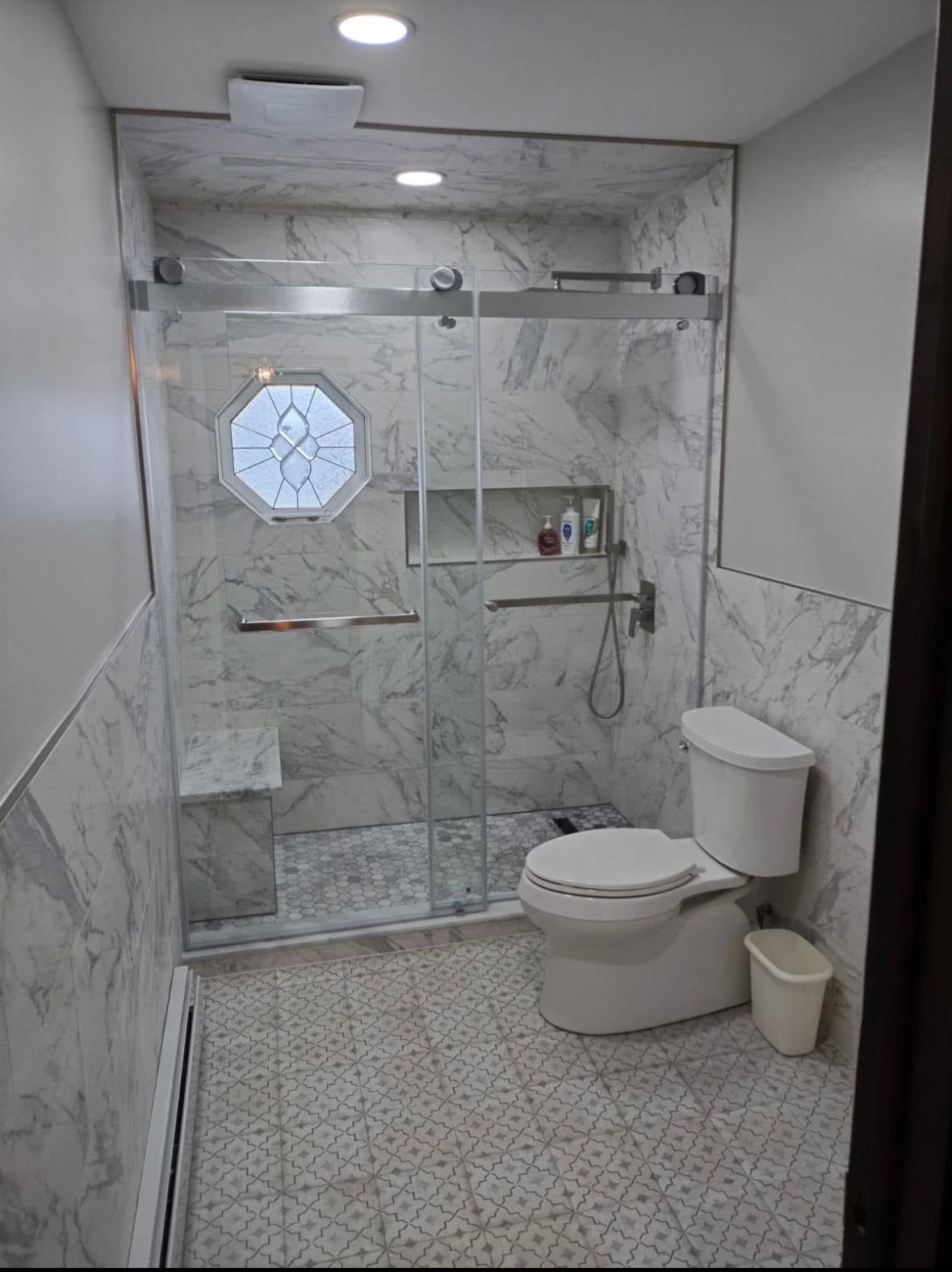 Small modern bathroom with marble tile, glass shower, toilet, and hexagonal window.