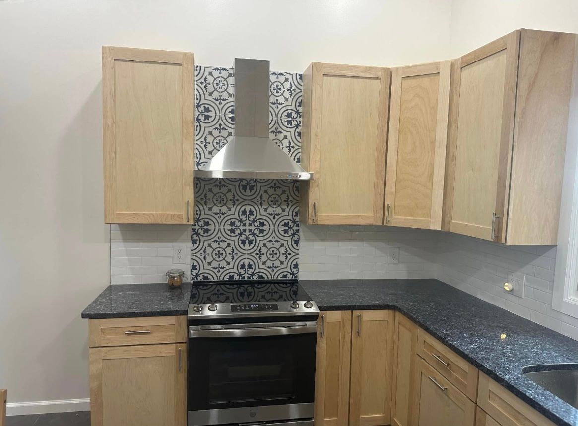 Small kitchen with light wood cabinets, black countertops, stainless stove, and patterned tile backsplash.