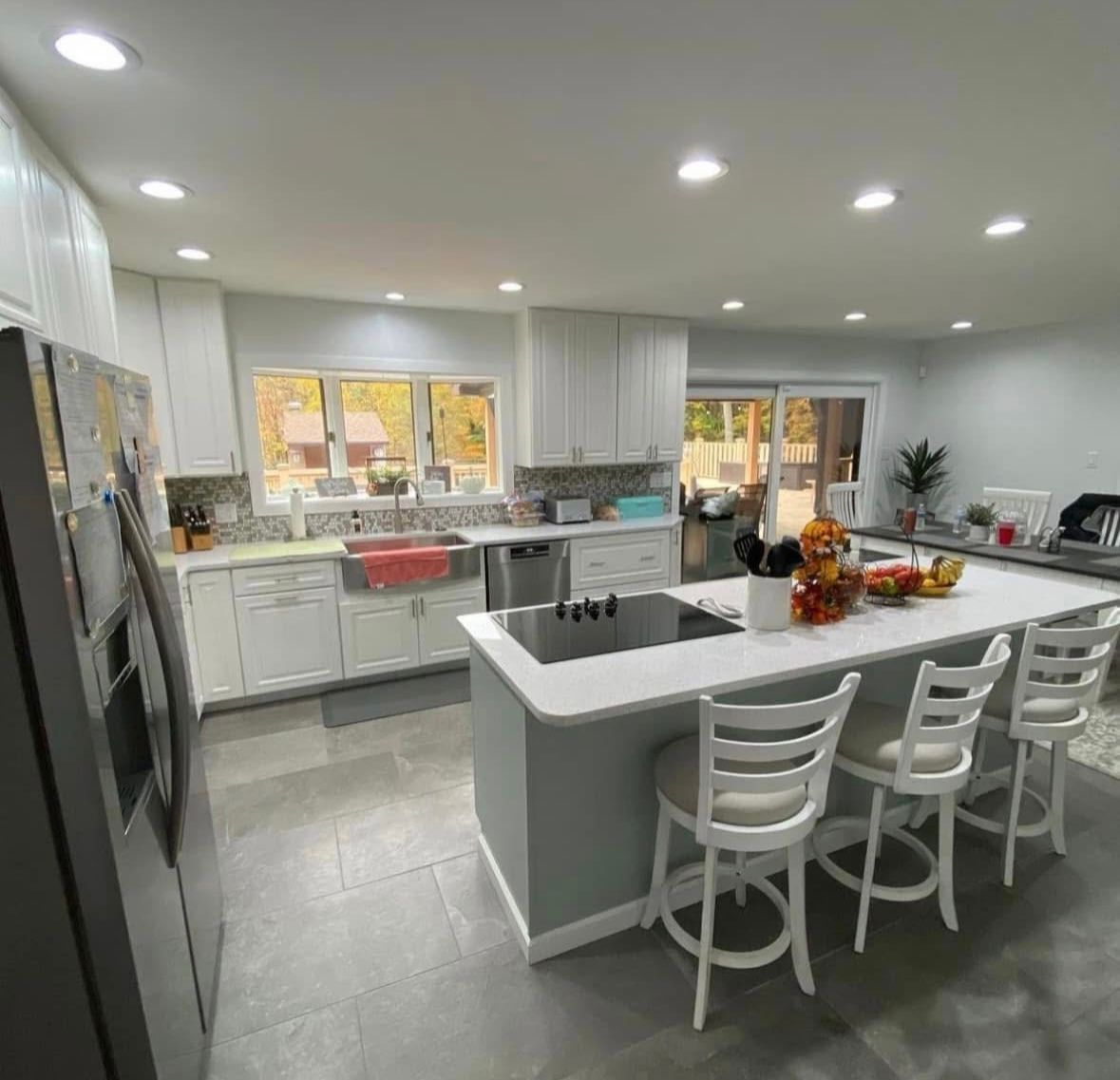 Bright modern kitchen with white cabinets, island seating, gray tile floor, and large windows showing autumn trees
