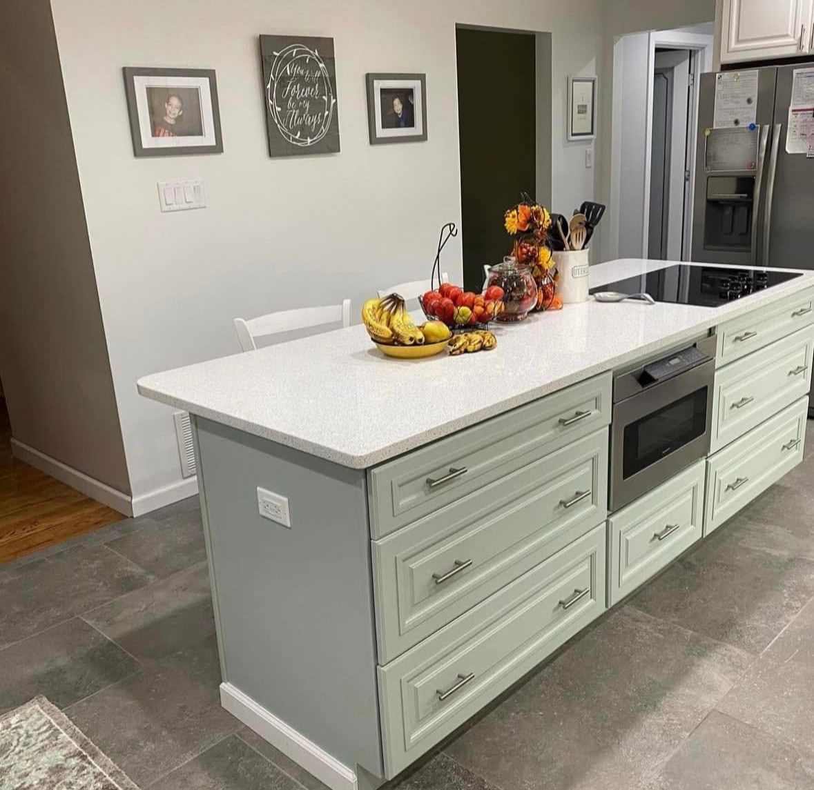 Modern kitchen with a large pale island, built-in oven, and fruit and flower centerpieces.