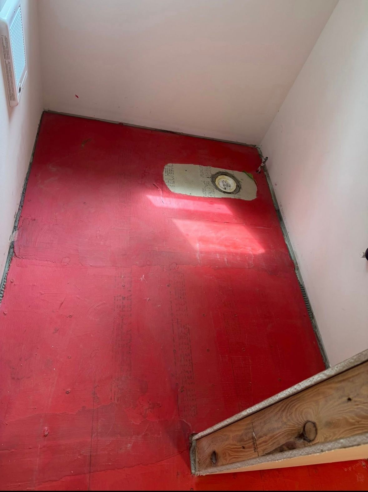 Small red-floored shower stall with a drain, white walls, and a wooden step at the entrance