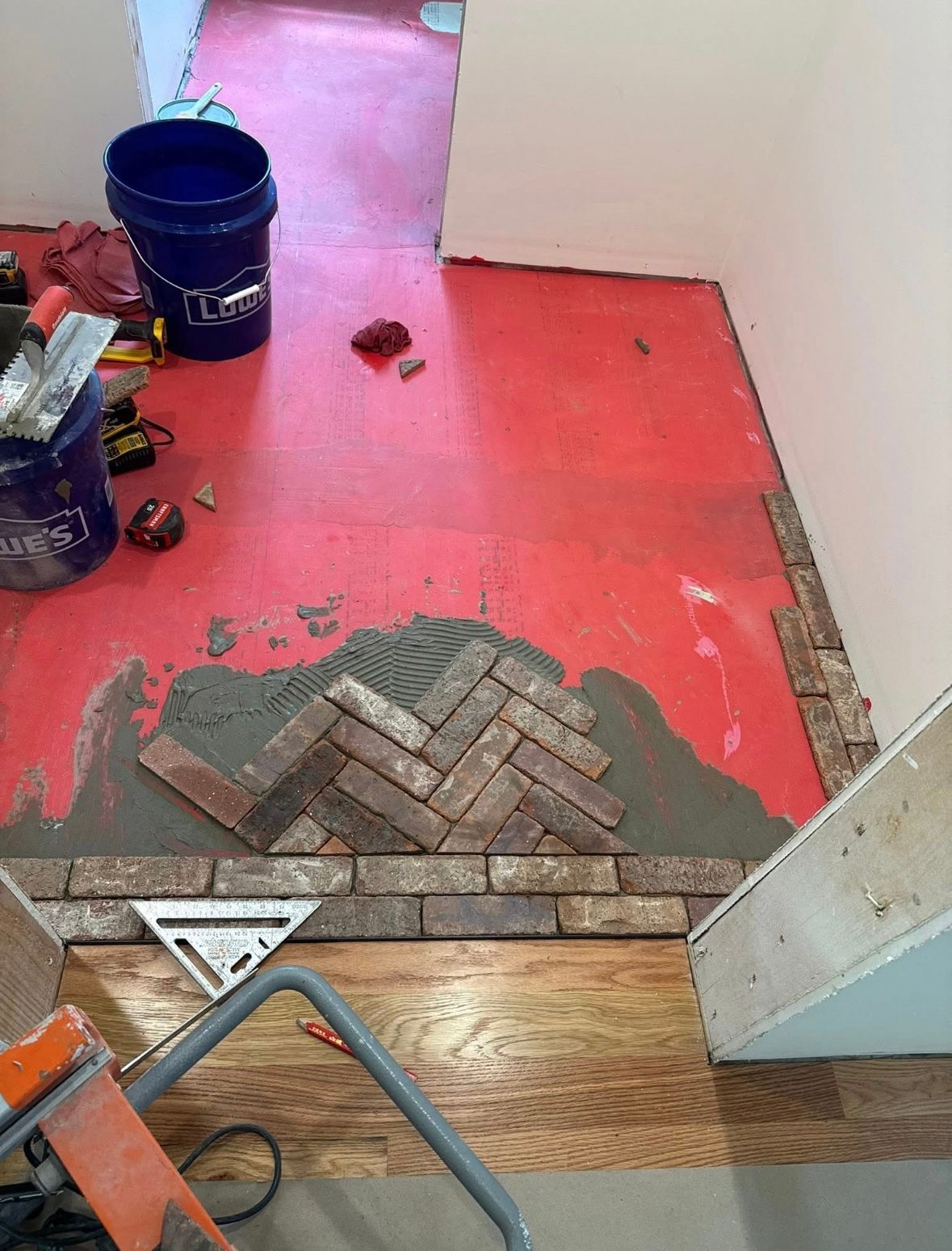 Partially tiled floor under renovation with red underlayment, buckets, tools, and a threshold transition