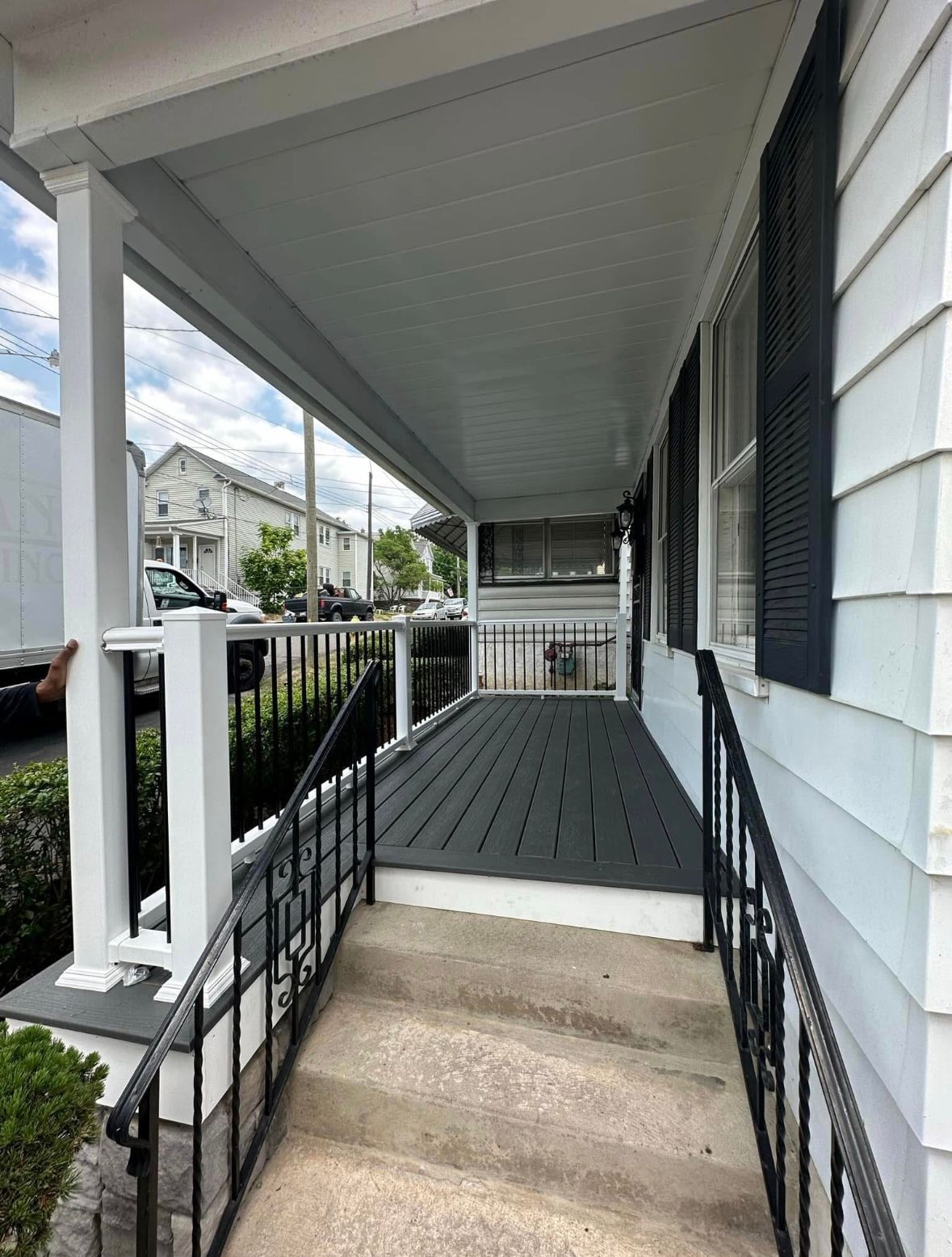 Covered front porch with white railings, gray flooring, and concrete steps leading up to a house entrance