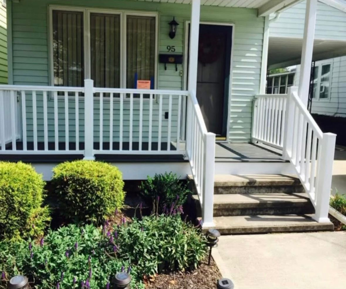 Front porch with white railings, steps, green siding, and neatly trimmed shrubs by the walkway