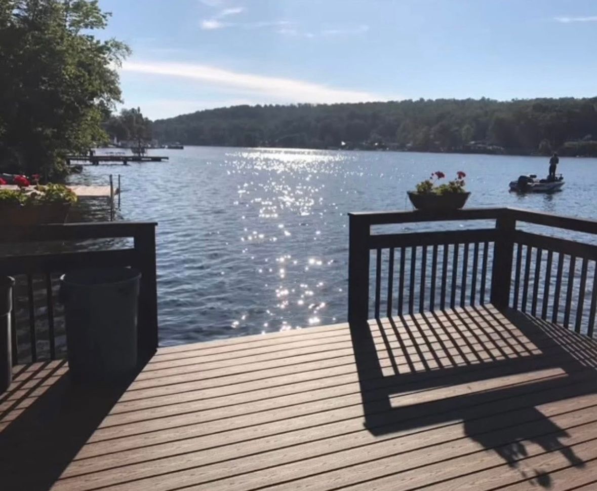 Wooden deck overlooking a sunlit lake with a small boat and tree-lined shore in the distance.
